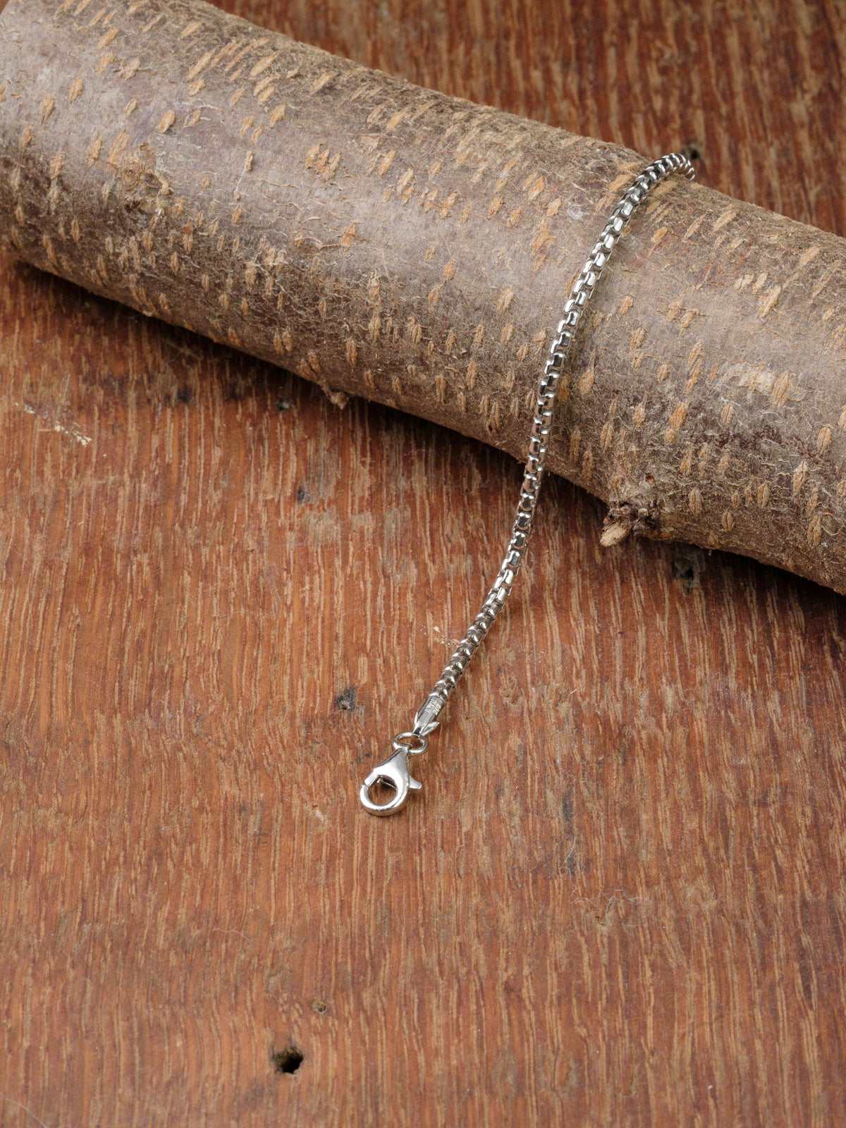 The Ezra 50 Bracelet by ID Menswear, crafted from 925 sterling silver with a lobster clasp, is shown on a textured wooden surface, accented by a diagonally placed piece of tree bark in the background.