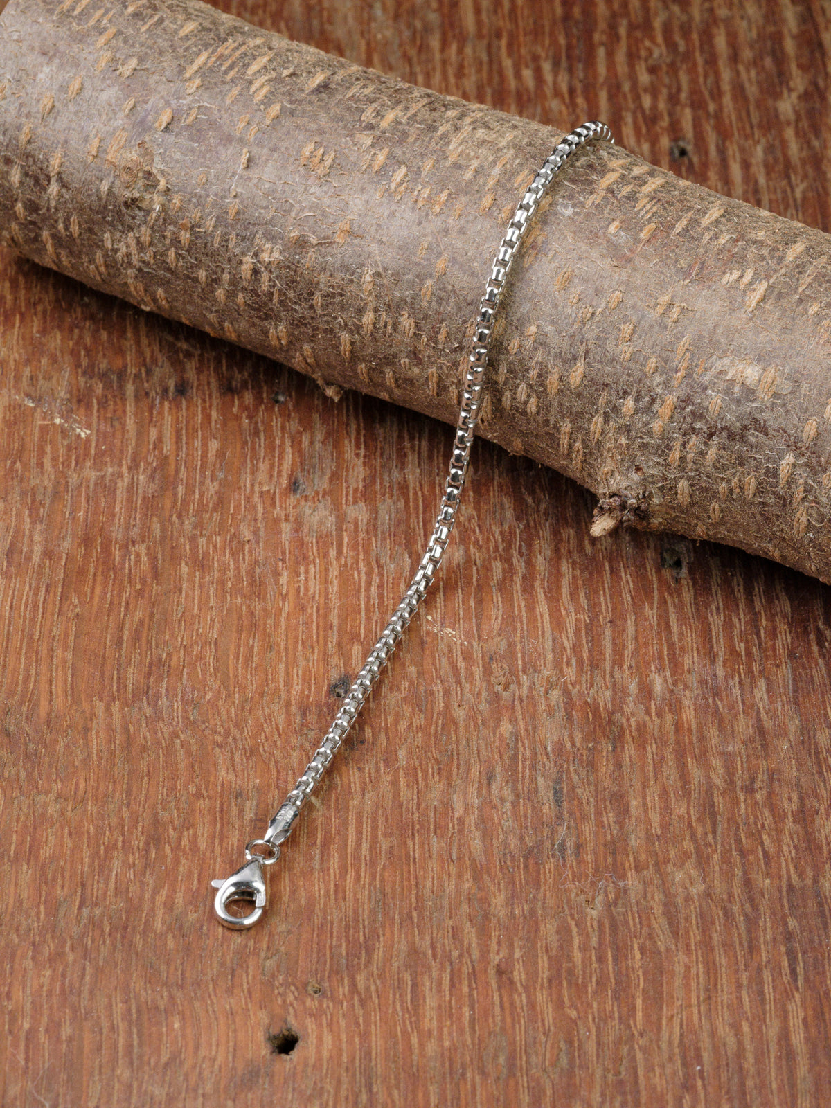 The Ezra 50 Bracelet by ID Menswear, crafted from 925 sterling silver with a box chain and lobster clasp, is displayed draped over textured, light-brown wood.