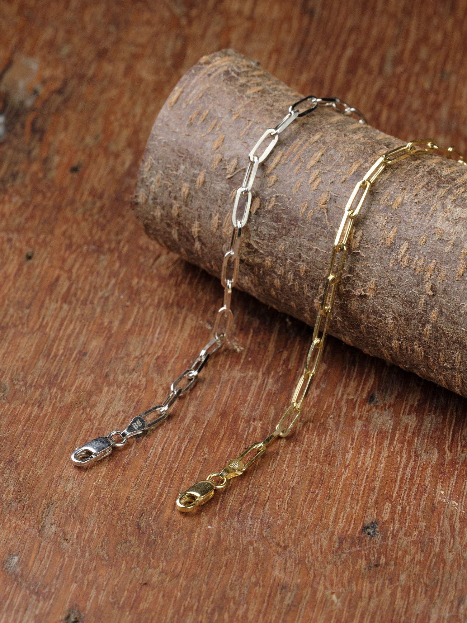 The Kai 100 Bracelet by ID Menswear, featuring an elongated paper clip chain and lobster clasp in both silver and gold tones, is displayed draped over textured wood on a rustic surface.