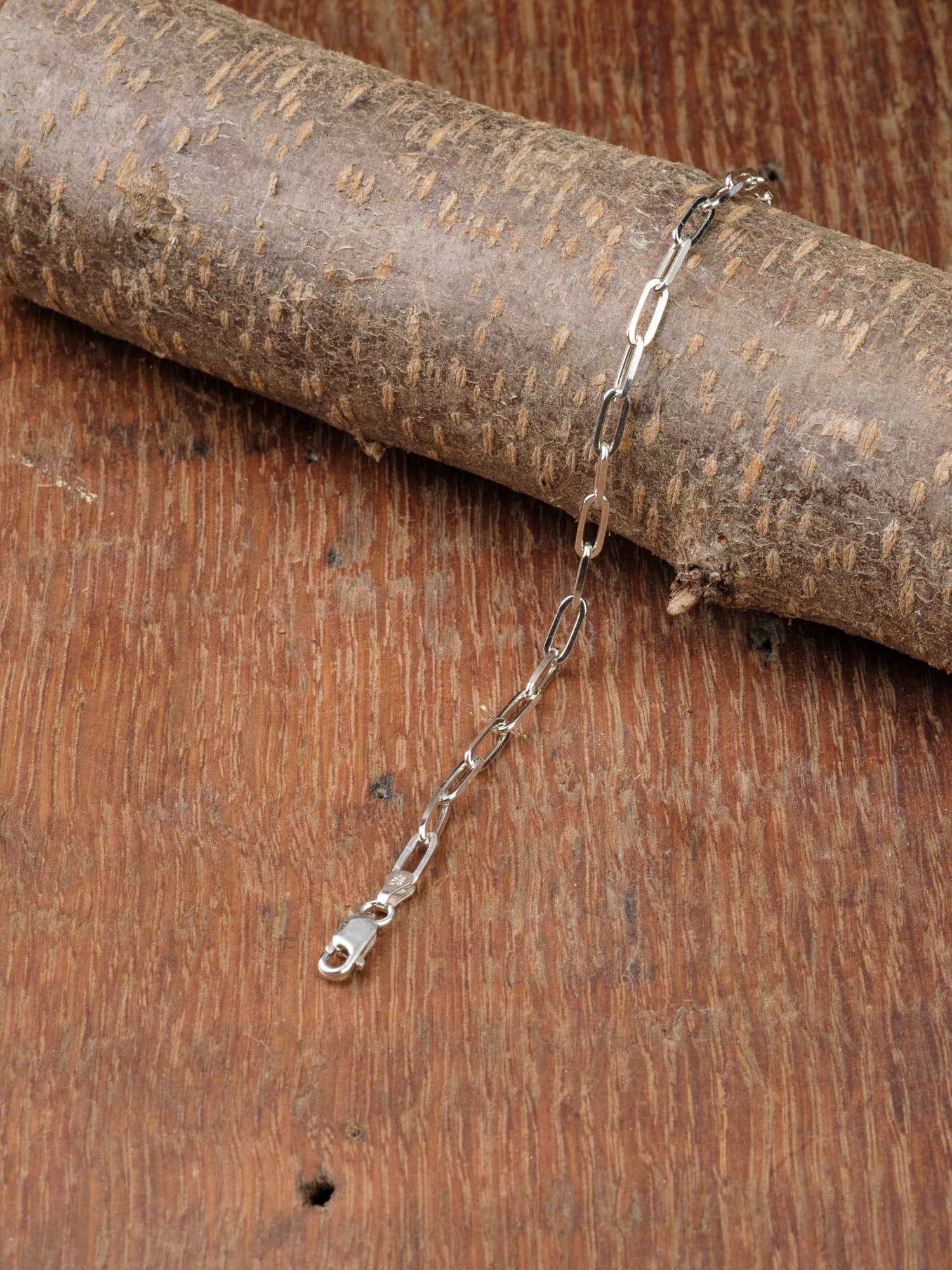 The Kai 100 Bracelet by ID Menswear, crafted in 925 Sterling Silver with elongated paper clip links and a clasp, is displayed on a rustic wooden surface and partially draped over a textured branch.