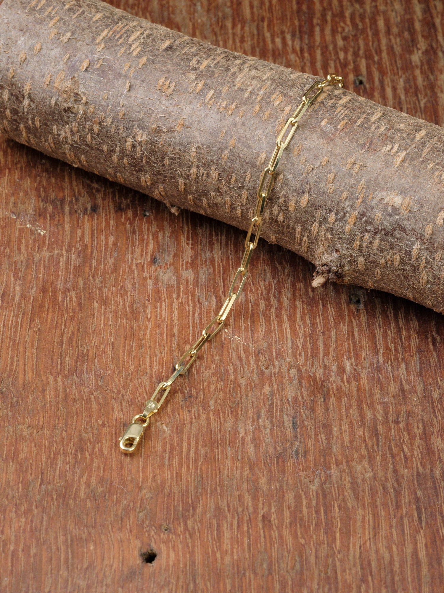 The Kai 100 Bracelet by ID Menswear, featuring delicate rectangular links, is displayed on a textured wooden surface and partially draped over a cylindrical piece of wood.