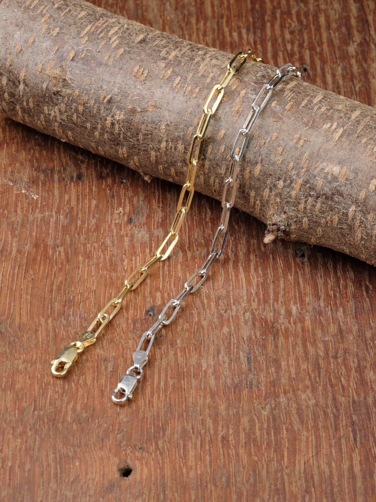 The Kai 120 Bracelets by ID Menswear—one gold plated, the other sterling silver—lie parallel on textured wood beside a log. Both feature elongated links and lobster claw clasps.