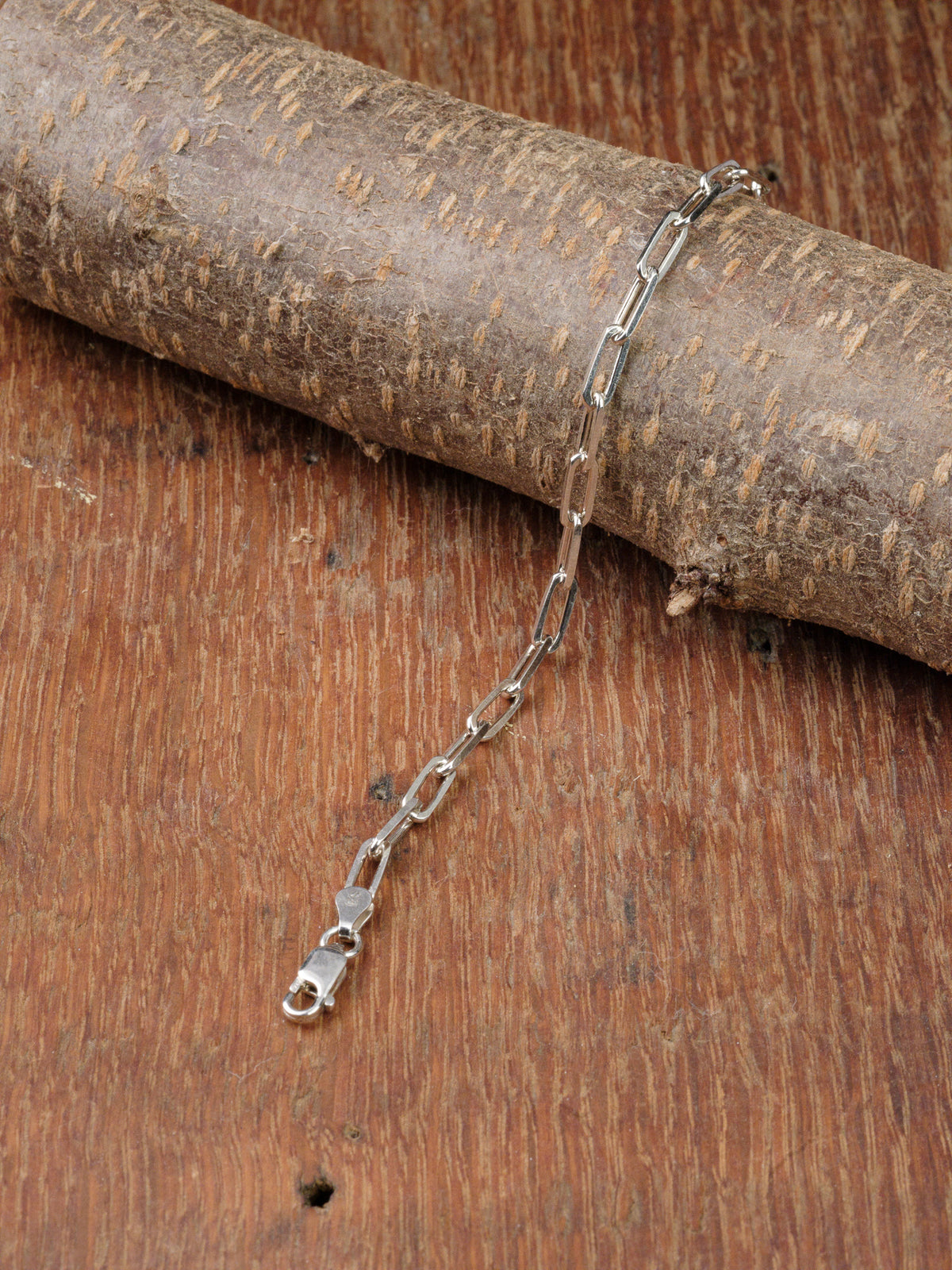 The Kai 120 Bracelet by ID Menswear, a sterling silver chain with rectangular links inspired by paper clip designs, is shown draped on a round piece of wood atop a textured wooden surface.