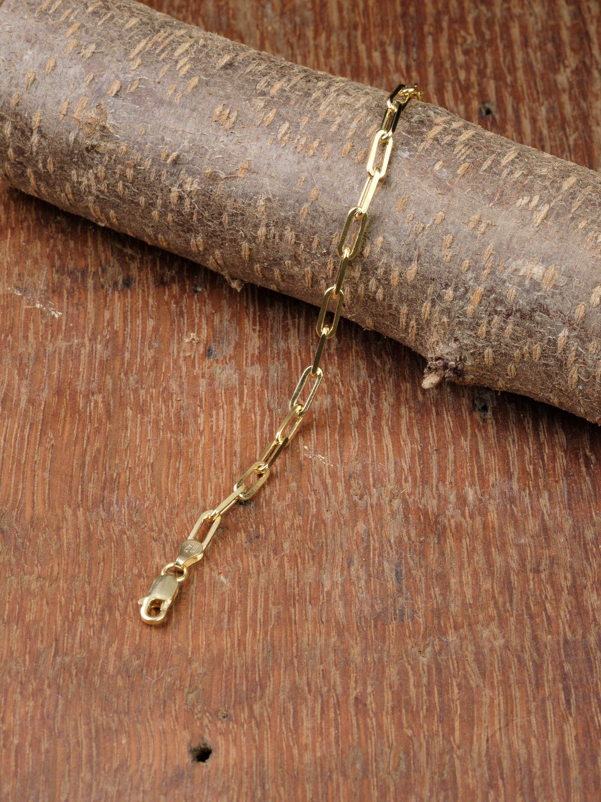 The Kai 120 Bracelet by ID Menswear, featuring gold-plated elongated links, is draped over a textured wooden branch on a rustic wooden surface.