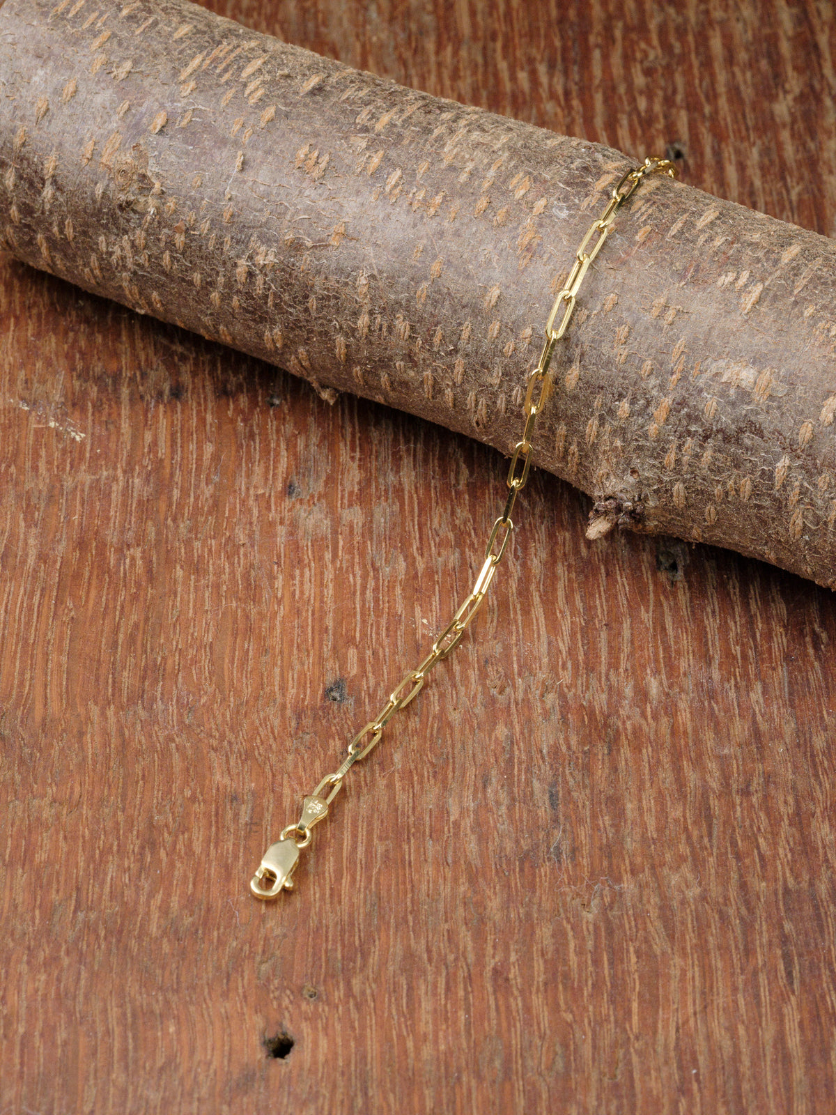 The Kai 80 Bracelet by ID Menswear, featuring an elongated gold-plated paperclip chain design, is displayed on textured wood set against a wooden background.