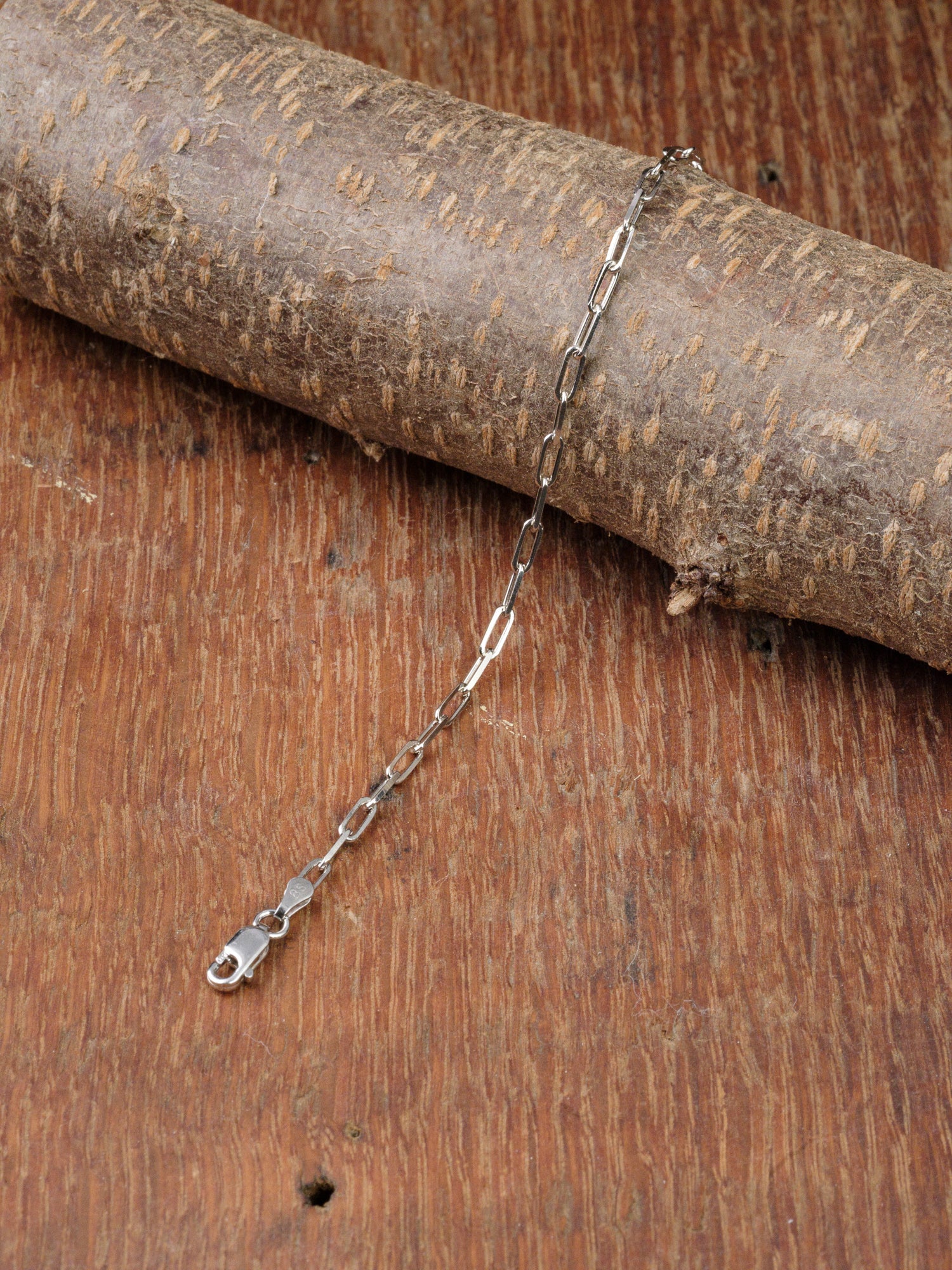 The Kai 80 Bracelet by ID Menswear, featuring a delicate sterling silver paper clip chain with elongated links, is showcased on a textured wooden surface and draped over a thick rustic branch.