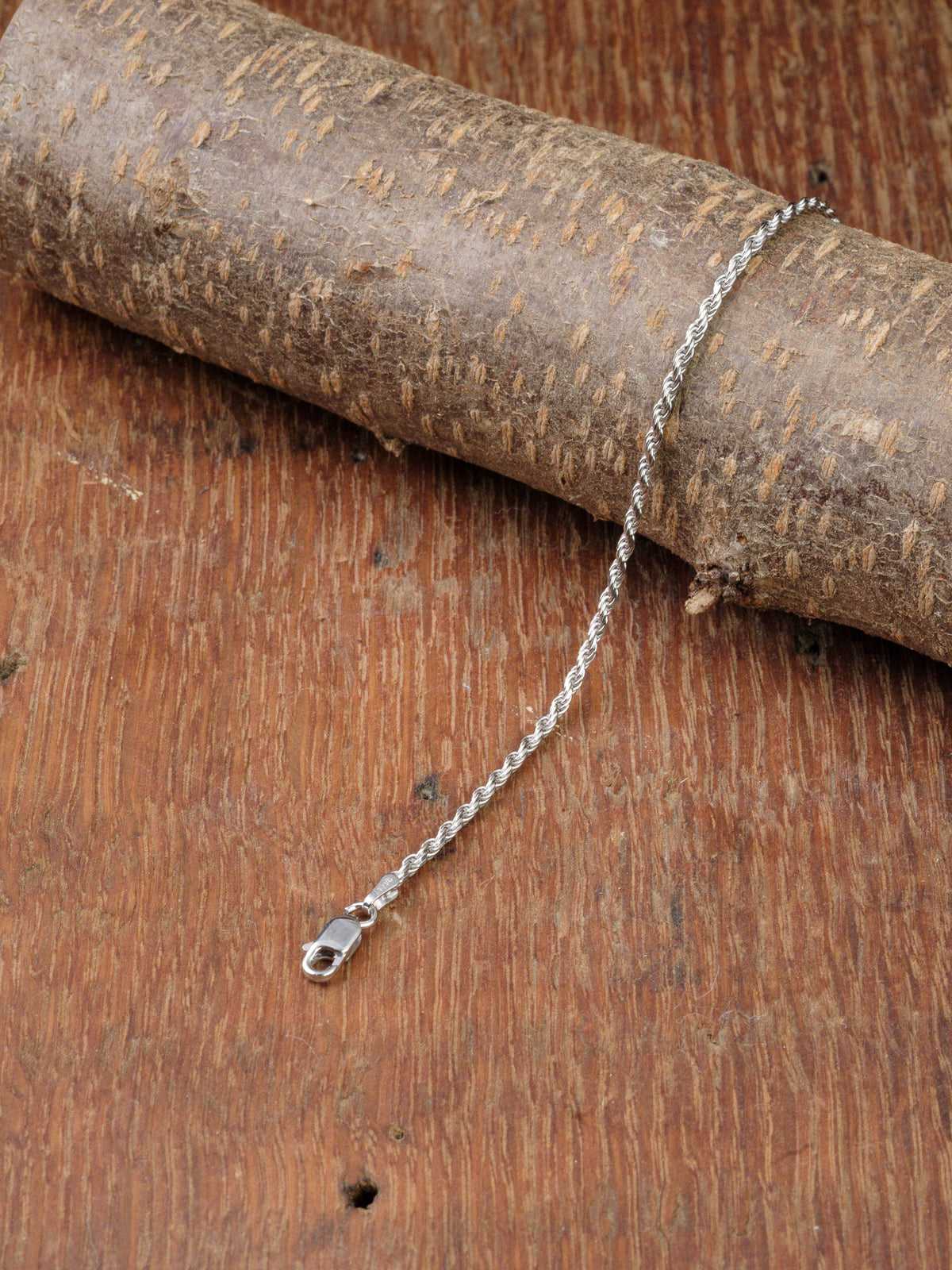 The Orion 40 Bracelet by ID Menswear, a slim 925 Sterling Silver Rope Chain with clasp, is shown draped over a cylindrical wood piece on a brown, textured wooden surface.