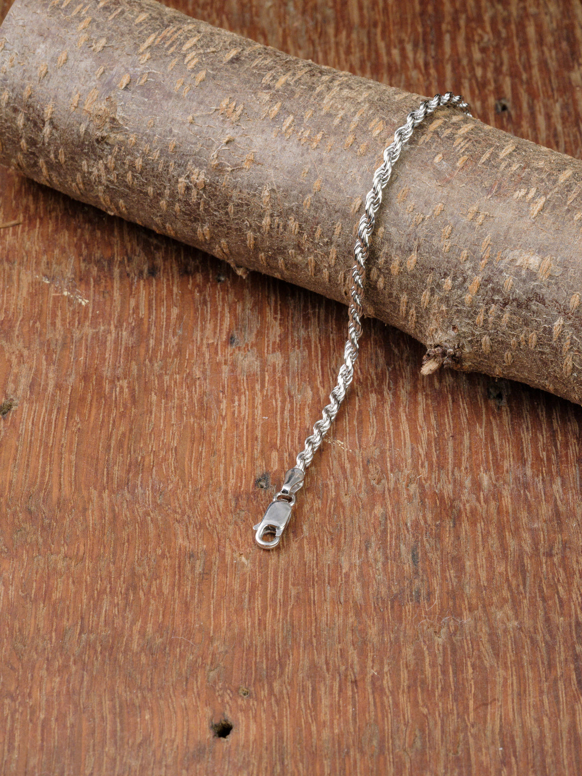 The Orion 50 Bracelet by ID Menswear, a 925 sterling silver rope chain with clasp, is elegantly displayed draped over tree bark on a textured wooden surface.