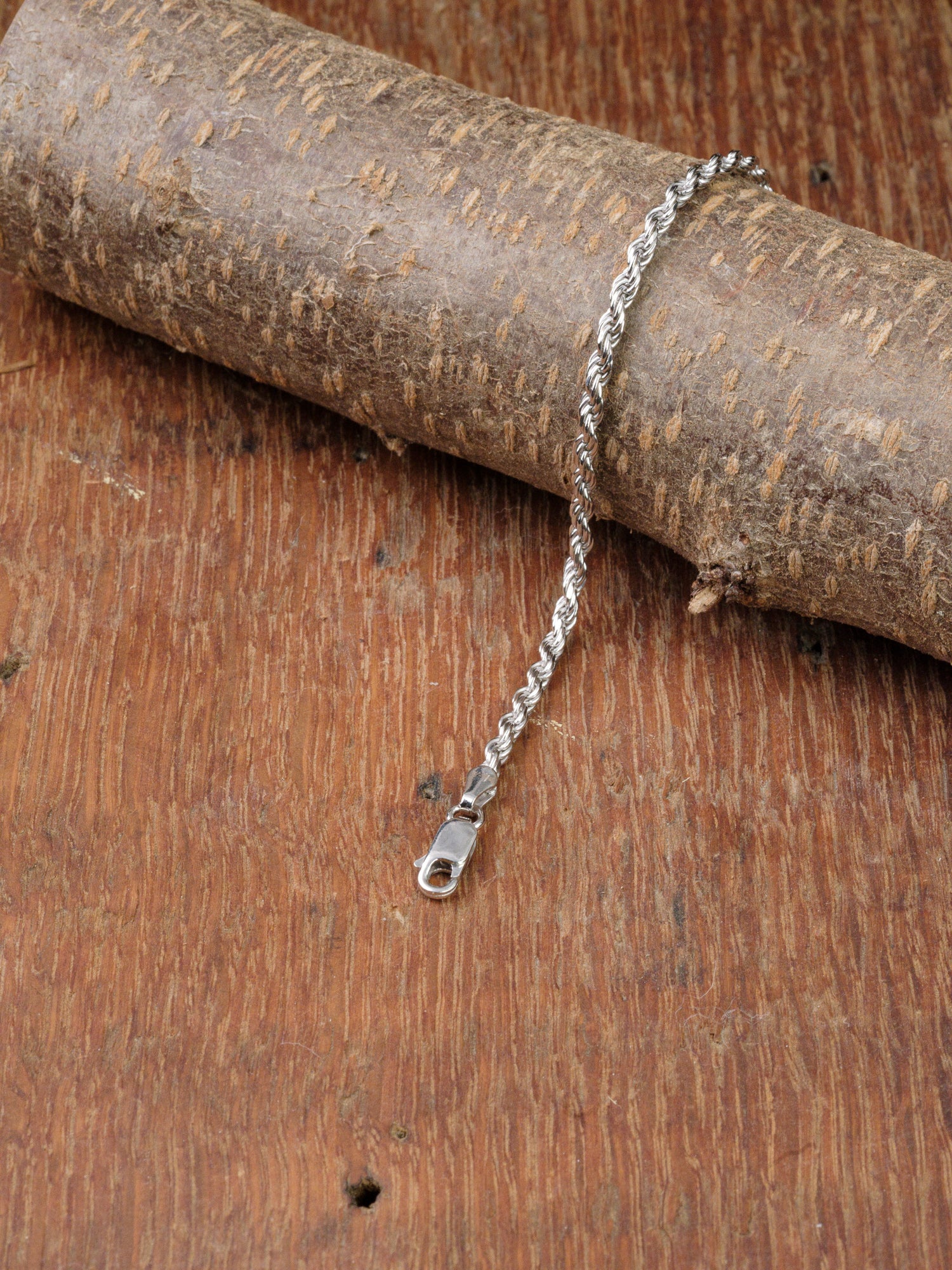 The Orion 50 Bracelet by ID Menswear, a 925 sterling silver rope chain with clasp, is elegantly displayed draped over tree bark on a textured wooden surface.