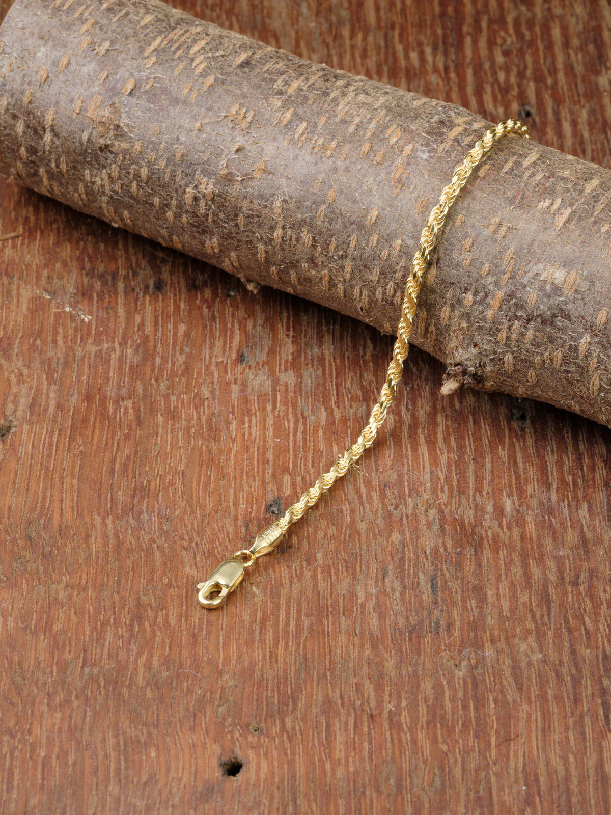 The Orion 50 Bracelet by ID Menswear, a gold-plated chain with a lobster clasp, is elegantly displayed on textured brown wood, adding timeless style to any look.