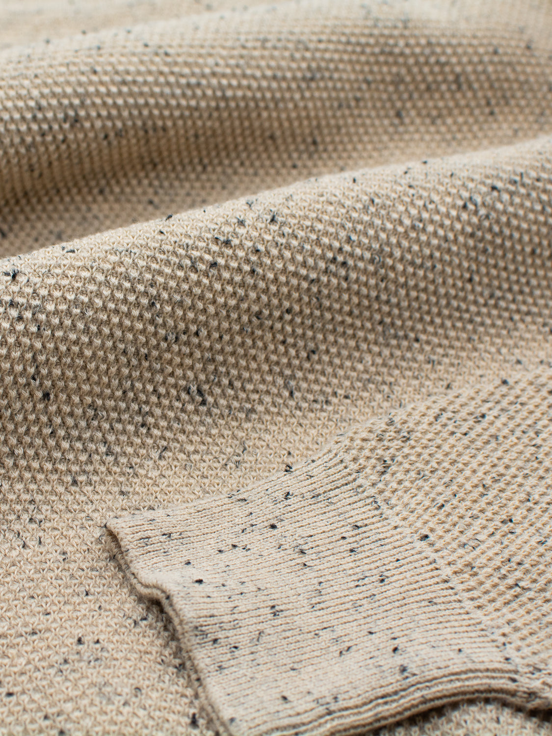 A close-up of beige knitted fabric with dark speckles from speckled yarn shows a textured pattern, highlighting the intricate weave and ribbed cuff. This image hints at ID Menswears Brampton High Neck Cotton Sweaters sophisticated style.