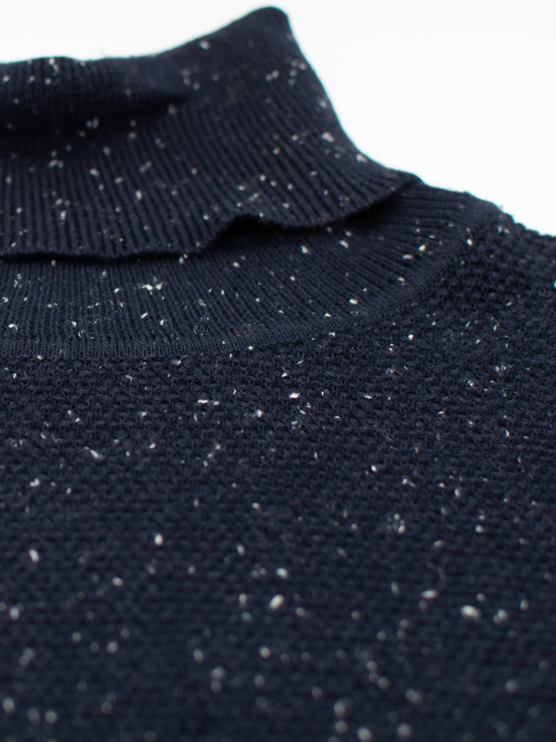Close-up of a dark blue Brampton High Neck Cotton Sweater by ID Menswear, showcasing a fine knit texture with white speckles. It features a high neck with the turtleneck collar folded to highlight the fabrics detail.