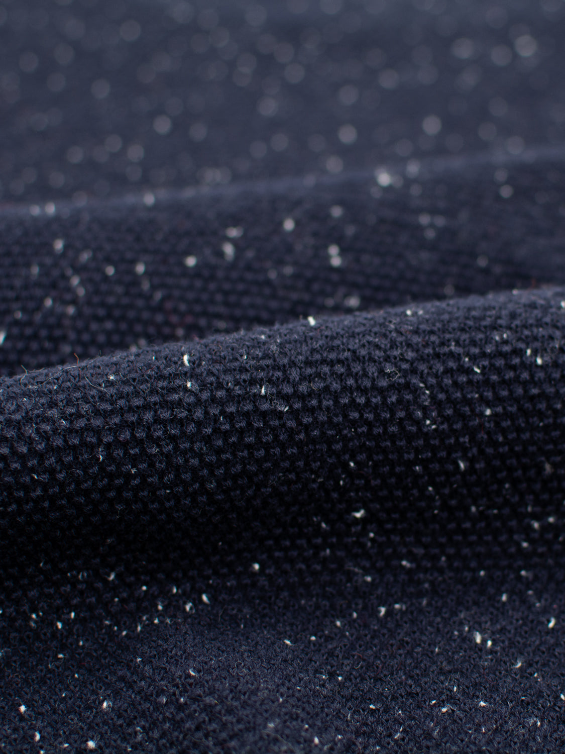 Close-up of a dark, textured fabric with small white specks from speckled yarn creates an intriguing look, forming gentle waves reminiscent of the intricate textures in the Brampton High Neck Cotton Sweater by ID Menswear.