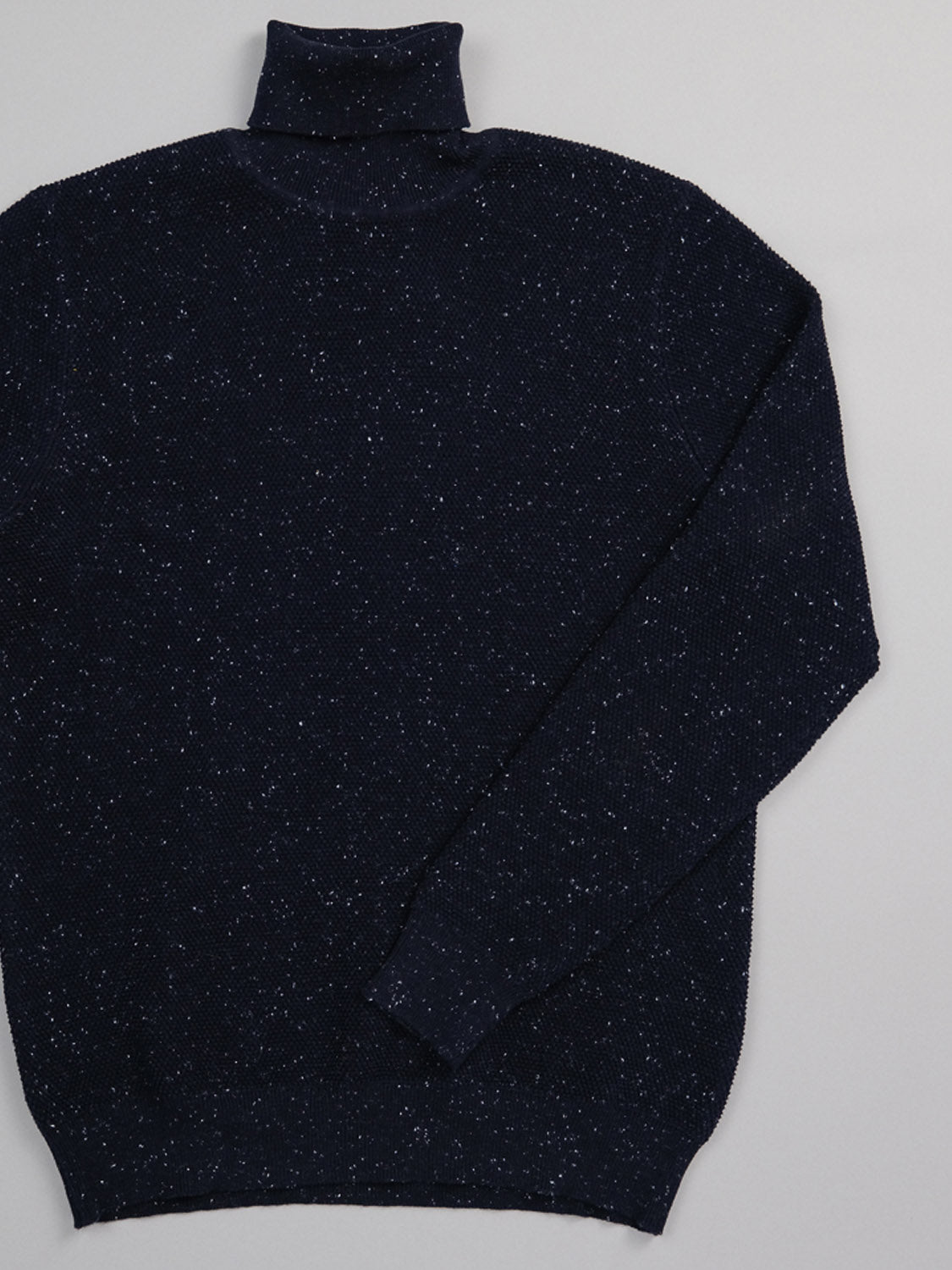 The Brampton High Neck Cotton Sweater by ID Menswear, made with speckled yarn, is displayed on a light gray surface. This dark navy turtleneck features a high neck and the right sleeve folded across, with small white specks providing a subtle contrast.