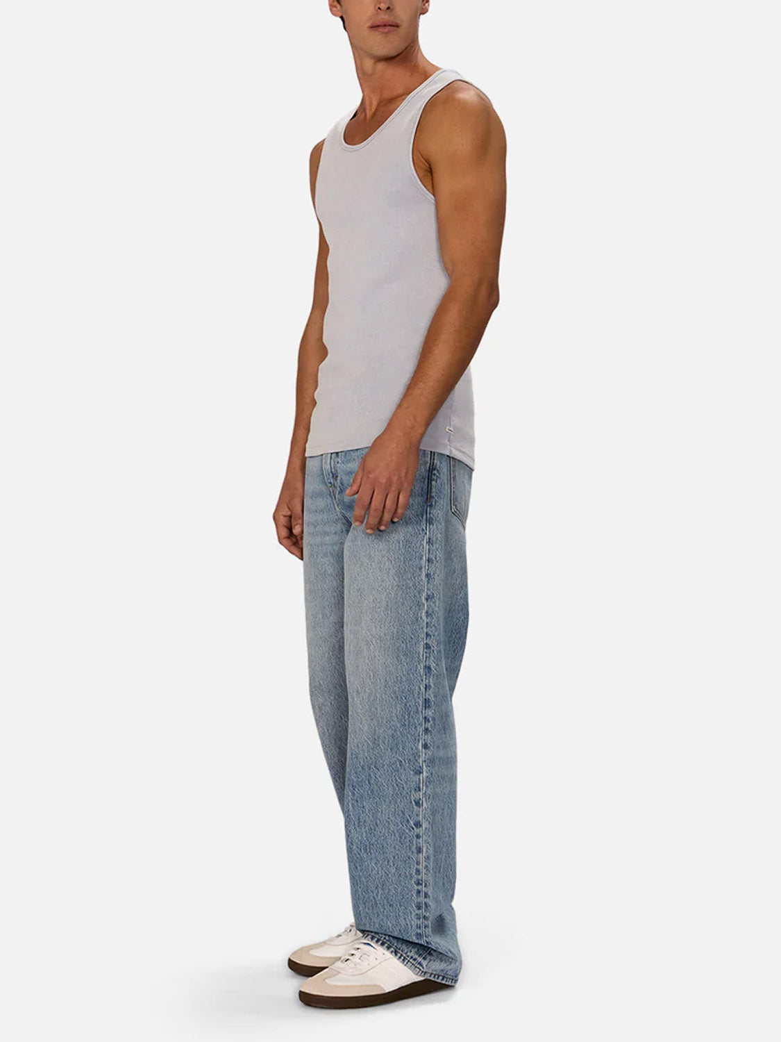 A person stands against a plain background, wearing Industrie’s Washed Brando Ribbed Tank Top in white, styled with light blue baggy jeans and white sneakers—a relaxed summer staple. Their head is turned slightly, arms resting casually by their sides.
