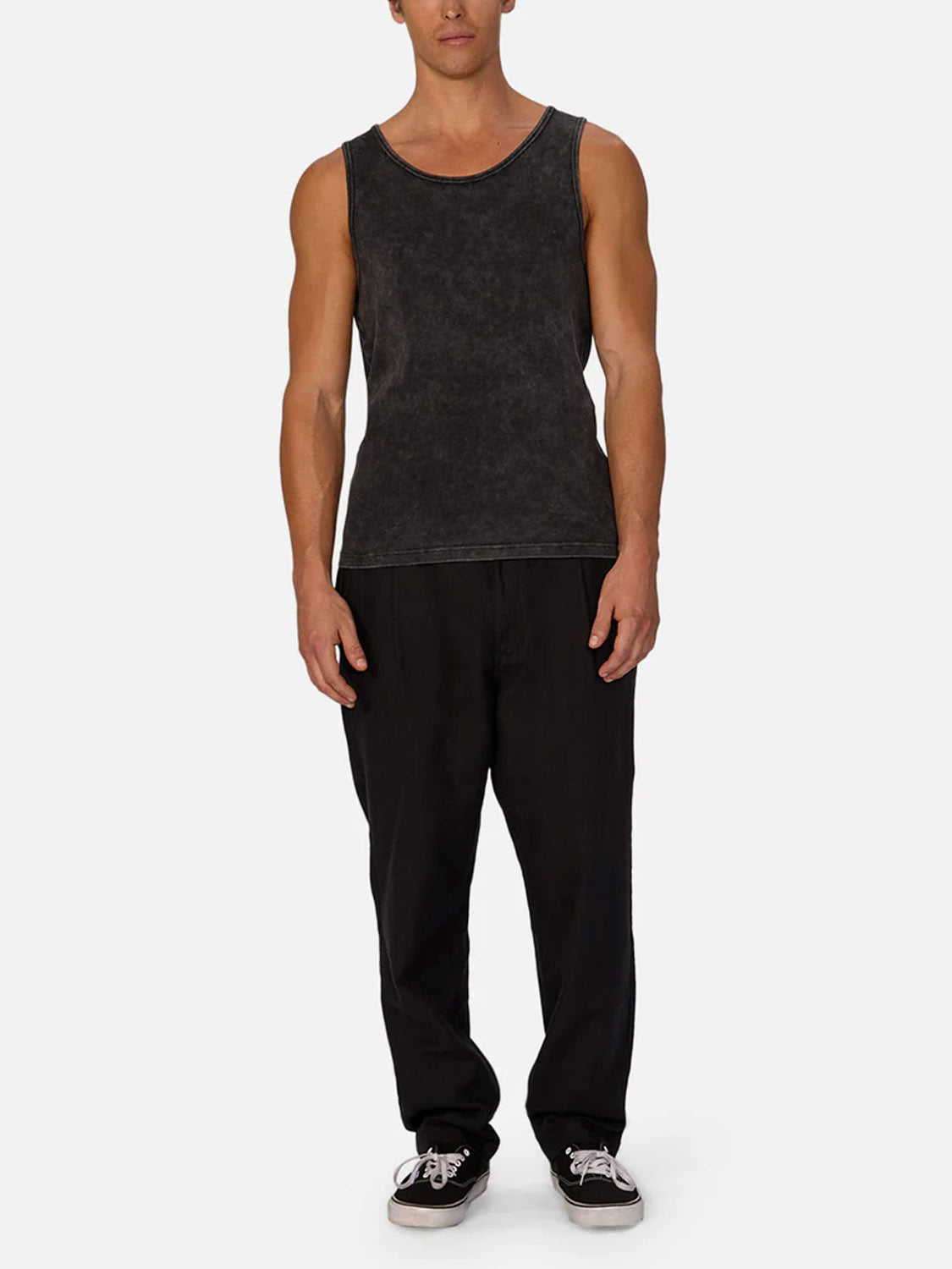 A man faces forward against a plain light background, wearing the Industrie Washed Brando Ribbed Tank Top—sleeveless and black—styled with loose black pants and black skate shoes with white soles for a summer-ready look.