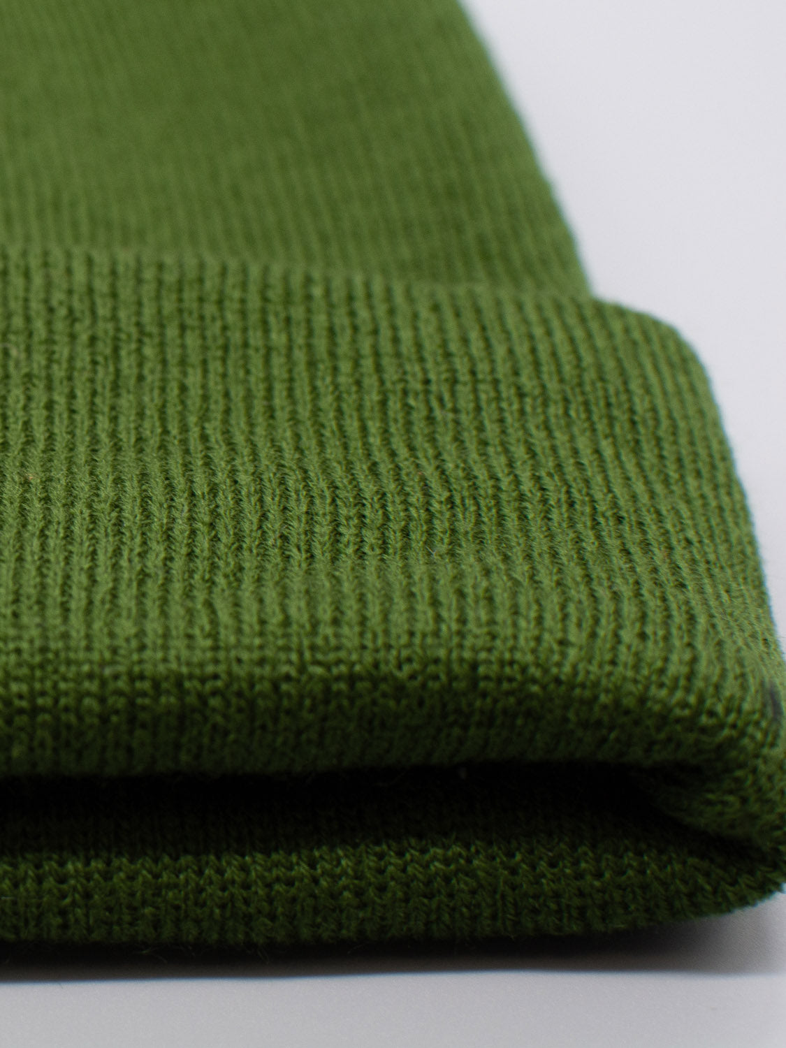 A close-up of the Bronx Acrylic Beanie by ID Menswear shows its green knit fabric with a folded edge, showcasing a fine knit pattern and ribbing. The lighting highlights its stretchable design and stylish color.