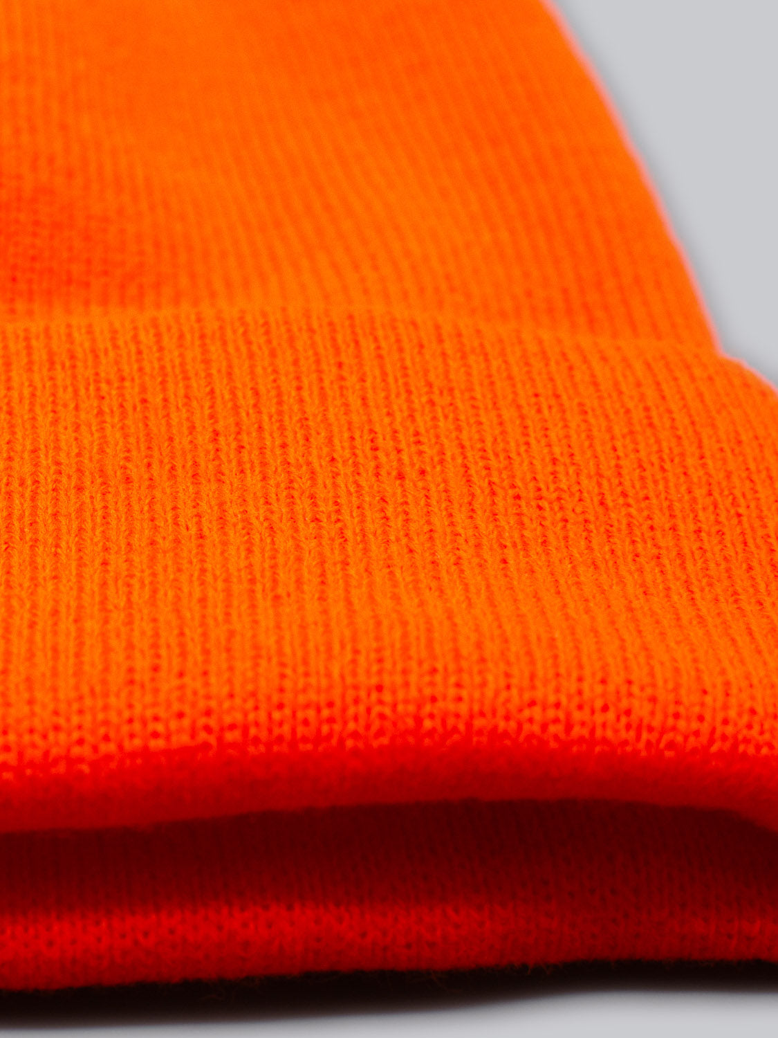 An up-close view of the Bronx Acrylic Beanie by ID Menswear, showcasing its bright orange acrylic knit texture and folded edge against a plain gray background, which highlights the beanies vivid color and stretchable design.