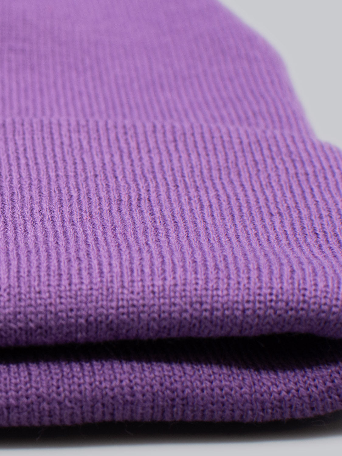 A close-up of a folded Bronx Acrylic Beanie by ID Menswear showcases its stretchable design and ribbed pattern. The soft, warm purple knit fabric is set against a neutral gray background.
