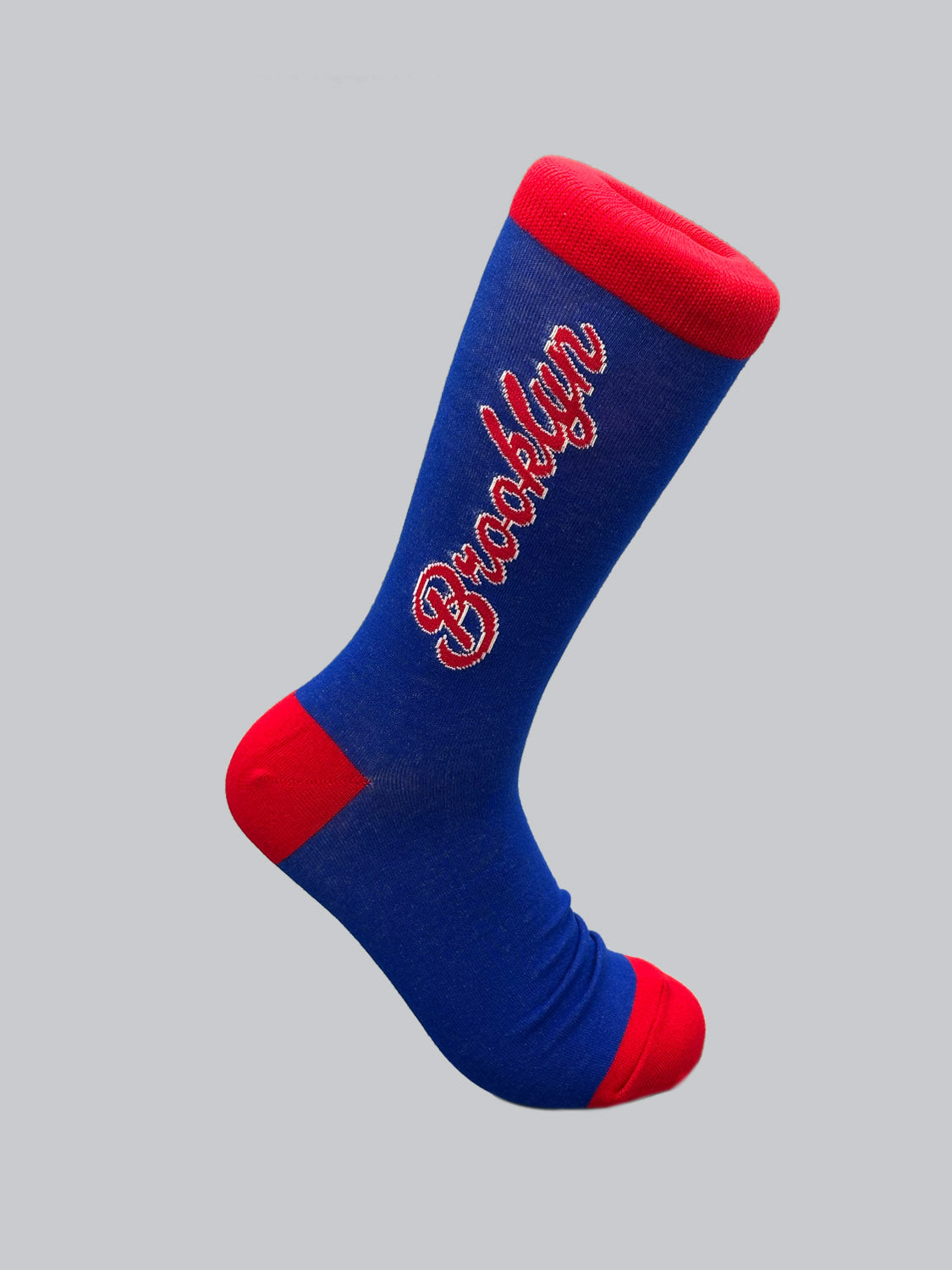 The Brooklyn Graphic Socks by ID Menswear are blue with red accents on the toe, heel, and cuff, featuring Brooklyn in red cursive. Made from premium combed cotton, they offer a versatile fit and are displayed on a plain gray background.