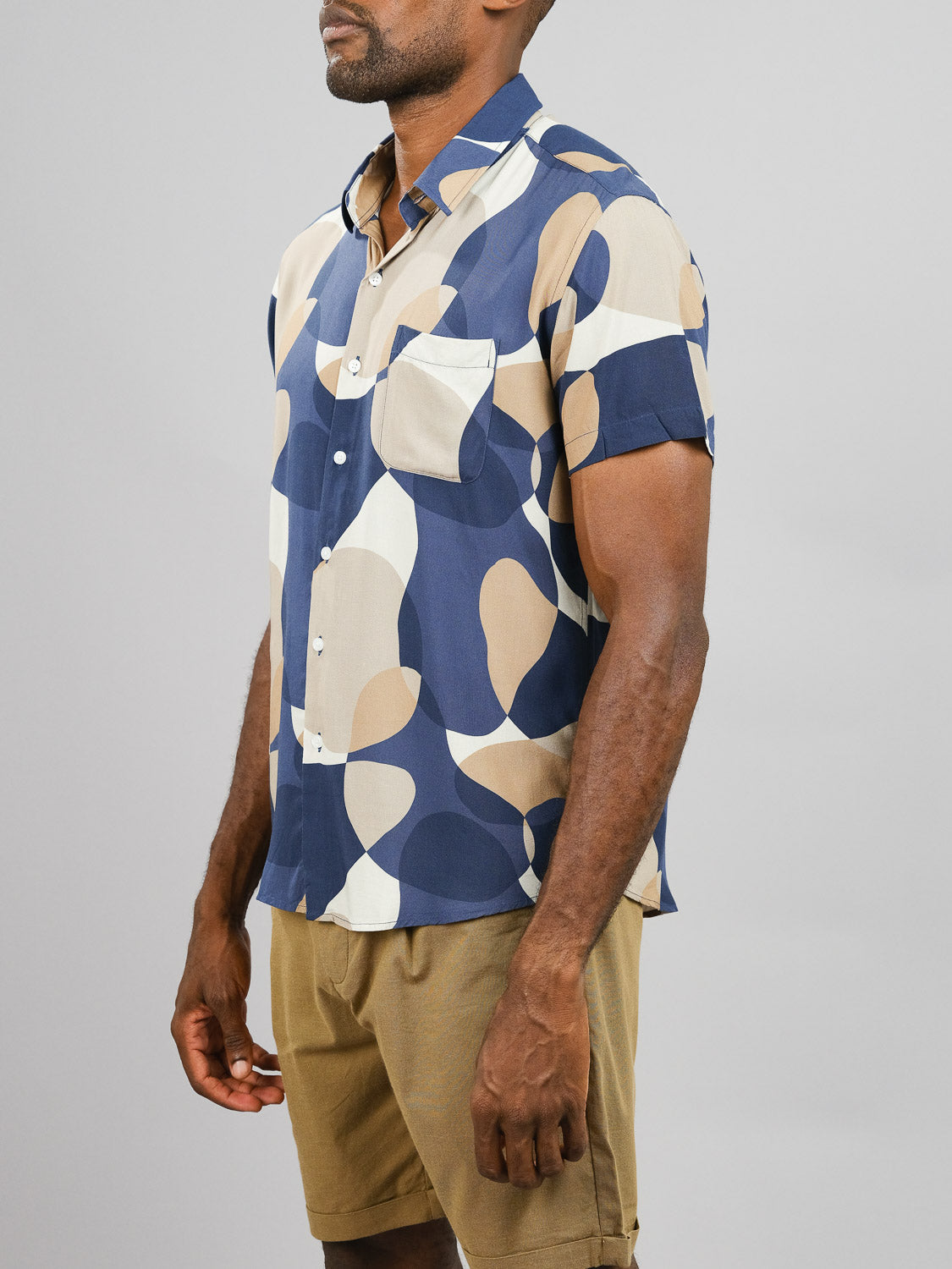 A man wears the ID Menswear Bubble Printed Rayon Shirt featuring blue, beige, and cream abstract patterns with tan shorts, posed at an angle against a plain light gray background.