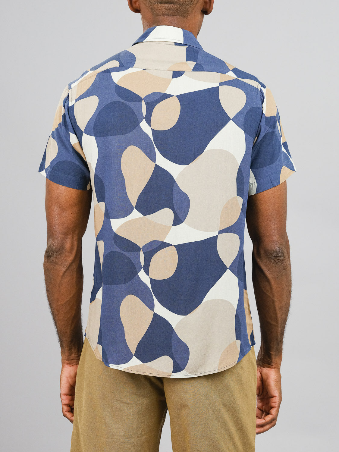 A man models the Bubble Printed Rayon Shirt by ID Menswear, showcasing its vibrant abstract blue, beige, and cream design with light brown pants against a gray background—ideal for warm weather style.