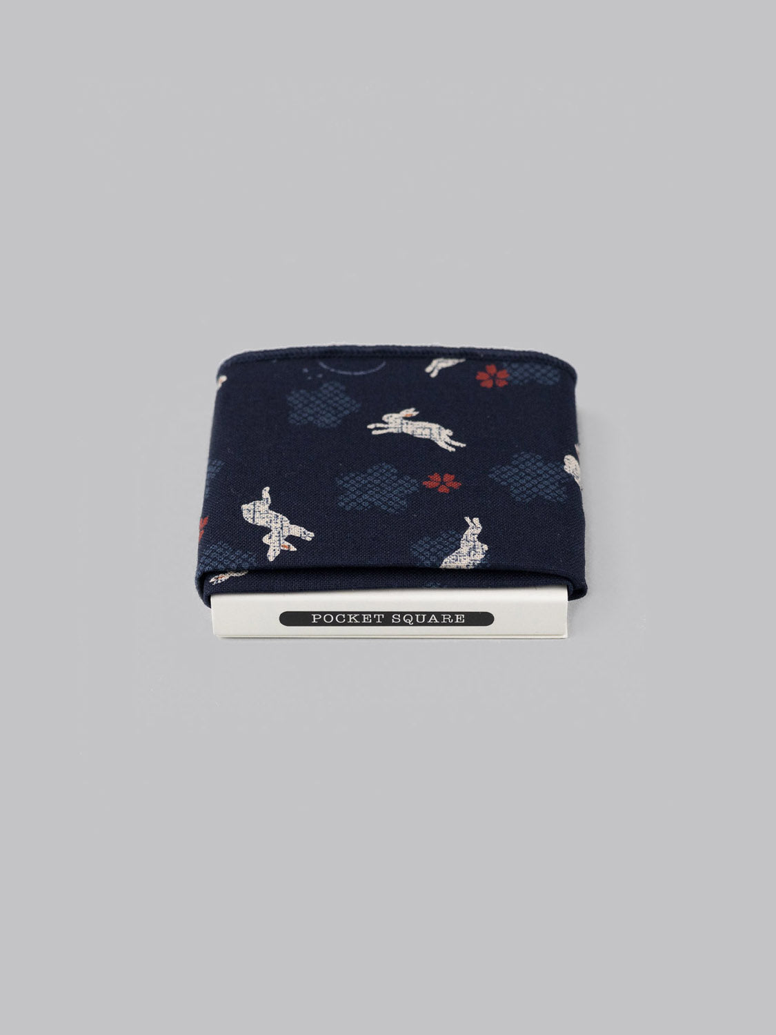 The Bunny Pocket Square by ID Menswear is a navy blue Japanese print pocket square with white rabbits and red-blue shapes, handmade in Brooklyn from 100% cotton, shown neatly folded in a white POCKET SQUARE holder against a grey background.