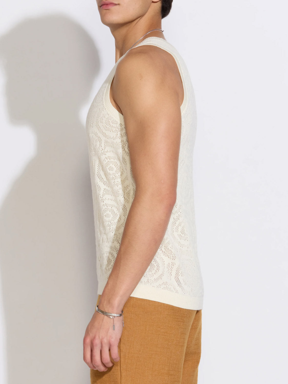 A person in profile wears the Civil Society Buyugo Sweater Knit Tank Top, a sleeveless cream 100% cotton piece, paired with tan pants, standing against a plain white background.