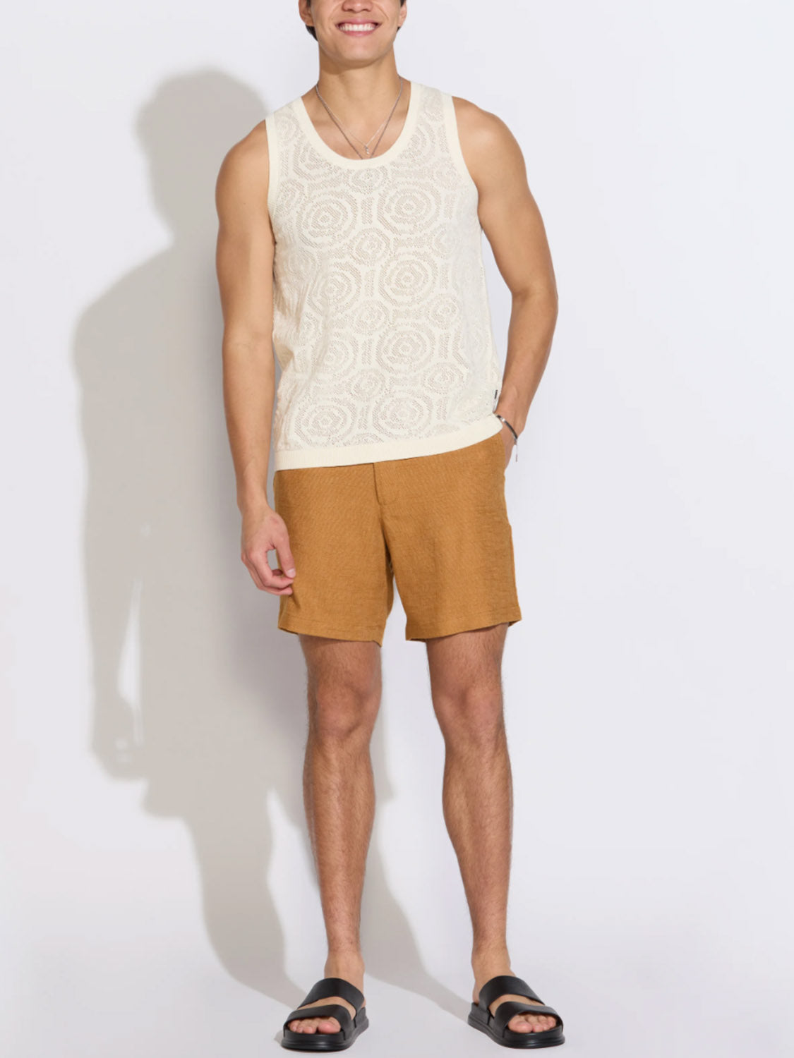 A man stands against a light background wearing the Civil Society Buyugo Sweater Knit Tank Top, light brown shorts, black slide sandals, and a necklace, with one hand in his pocket and a slight smile.