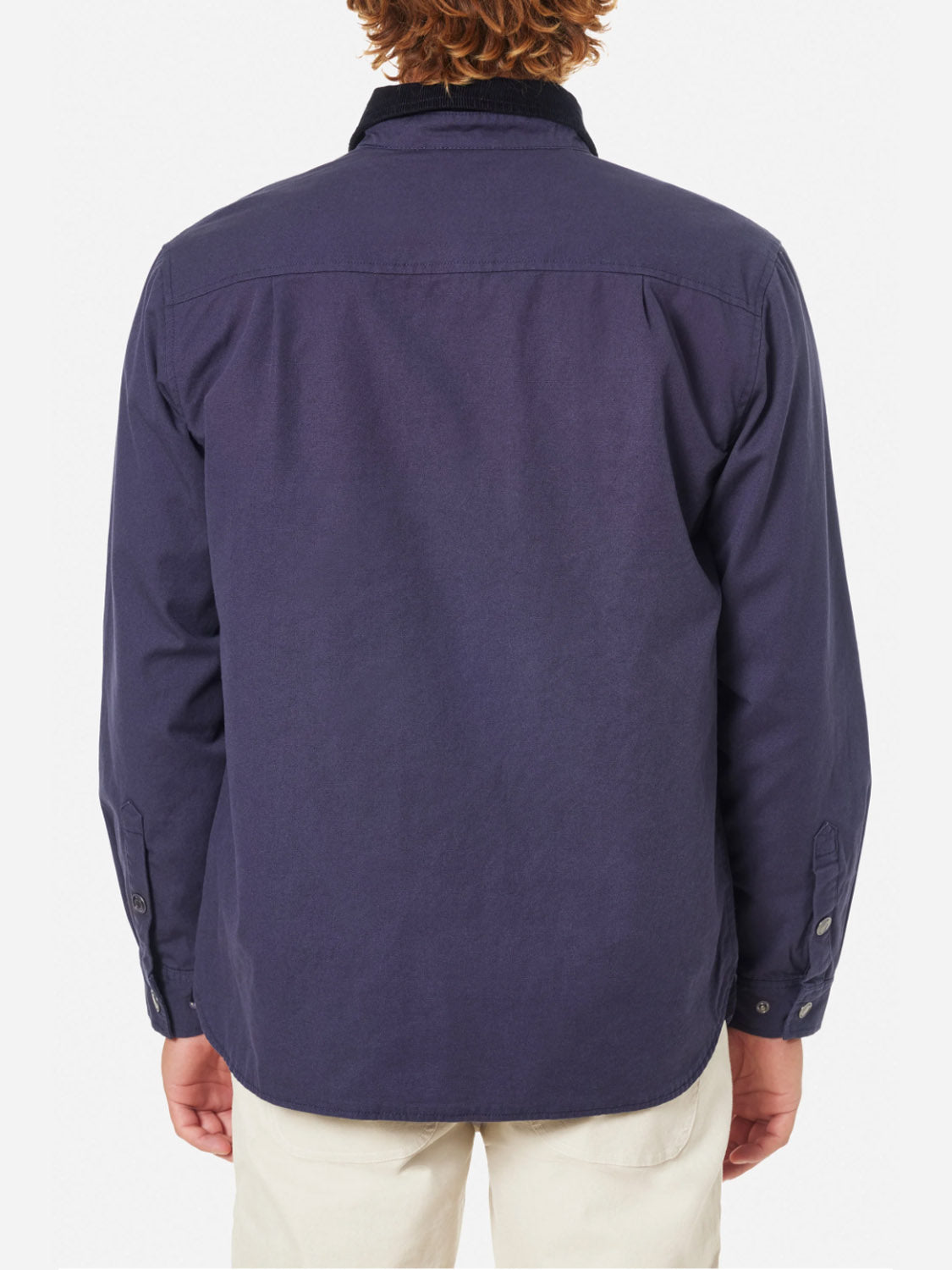 The image shows a person from behind wearing the Katin Campbell Jacket, featuring long sleeves and a corduroy collar. The dark blue jacket is untucked and paired with light-colored pants, while the persons curly hair is visible.