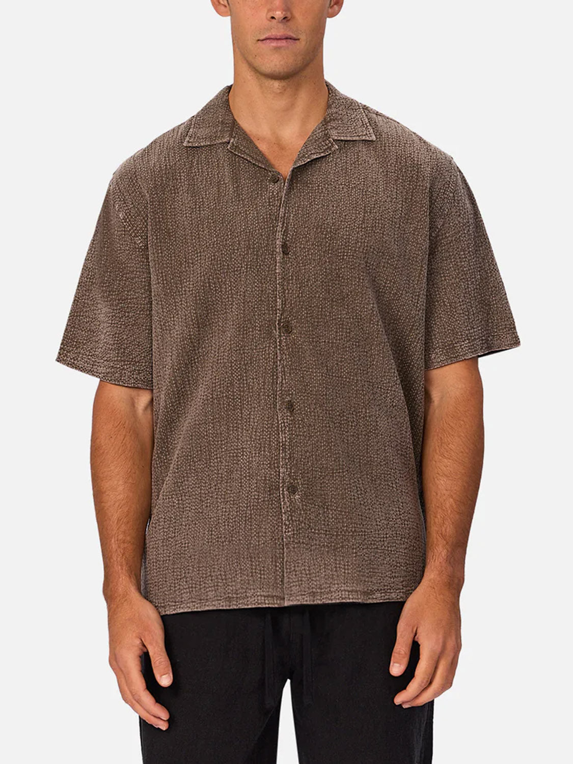 A man wearing Industrie’s The Canyon Short Sleeve Shirt in textured brown cheesecloth with a relaxed fit and black pants stands against a plain white background.