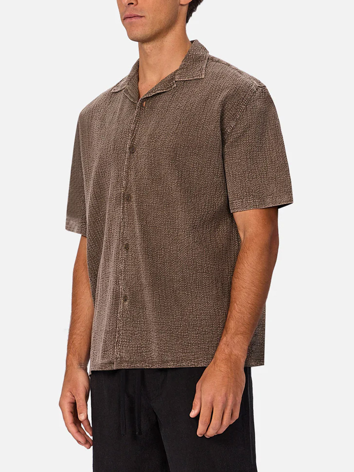 A man wearing Industrie’s The Canyon Short Sleeve Shirt in brown cheesecloth with a collar and black pants, standing against a plain white background.