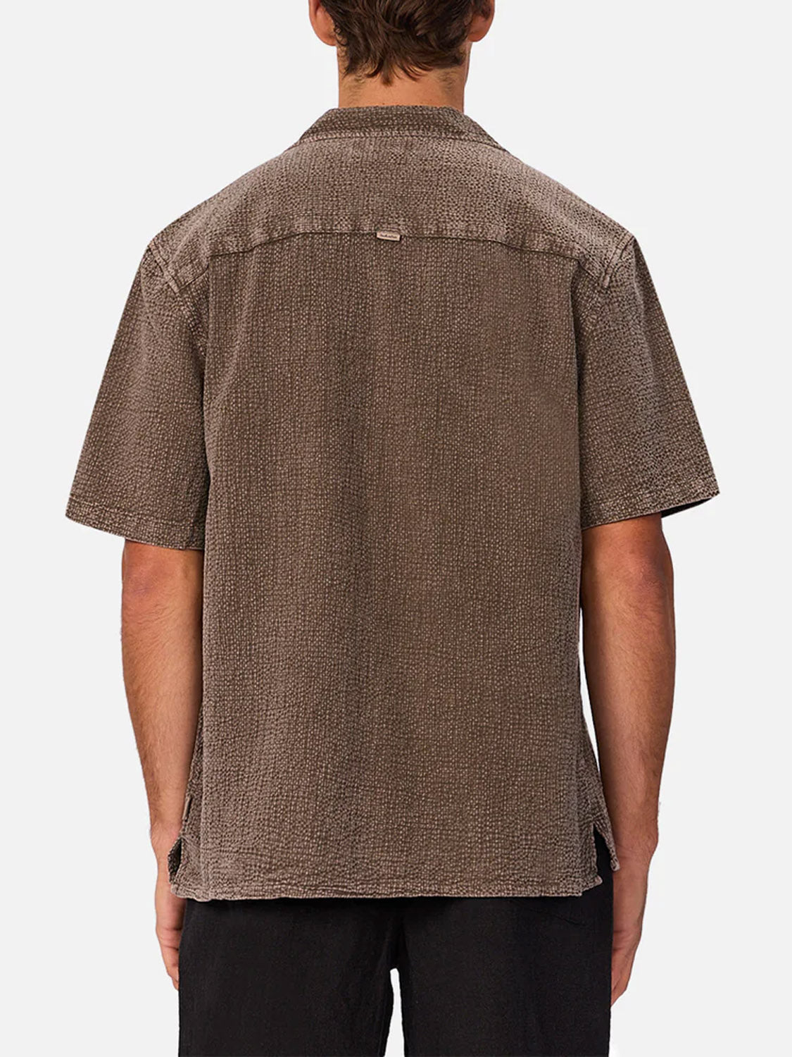 A person is seen from the back wearing Industrie’s The Canyon Short Sleeve Shirt in textured brown cheesecloth and black pants, set against a plain white background.