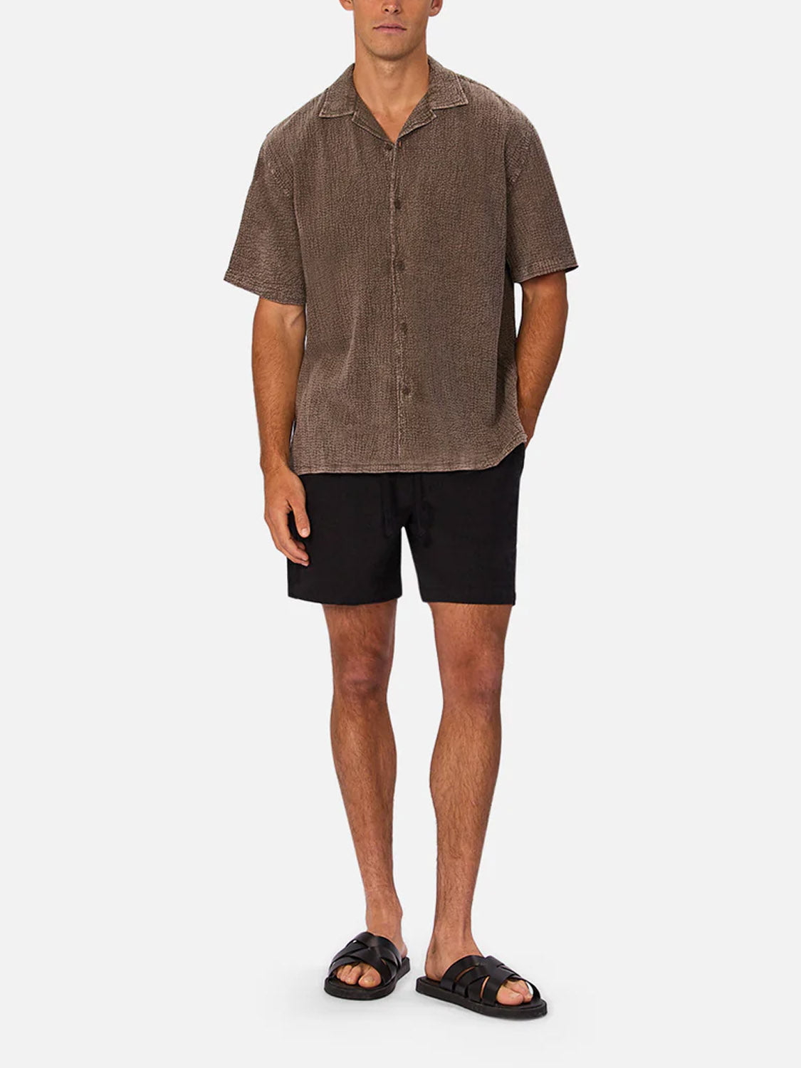 A man stands against a plain background wearing Industrie’s The Canyon Short Sleeve Shirt in textured brown, paired with black shorts and black slide sandals. His arms rest relaxed at his sides.