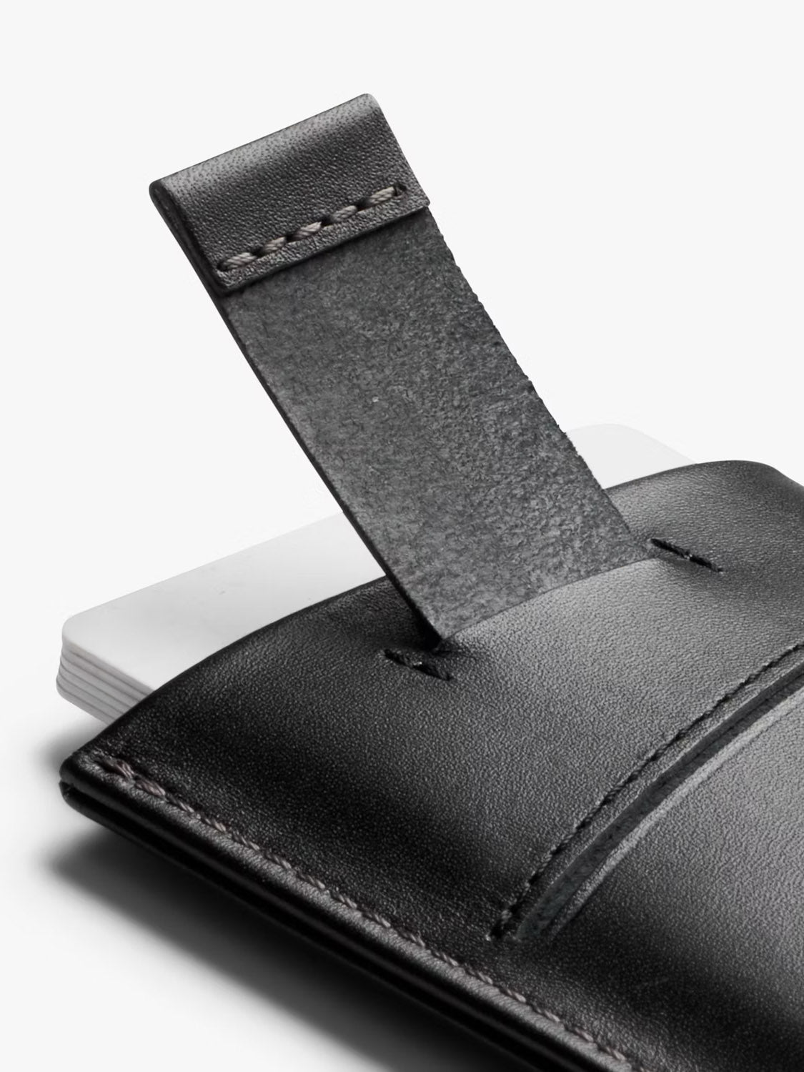 A close-up of the Bellroy Card Sleeve Wallet in black leather features a partially raised pull-tab, exposing the edges of several white cards tucked inside the main pocket.