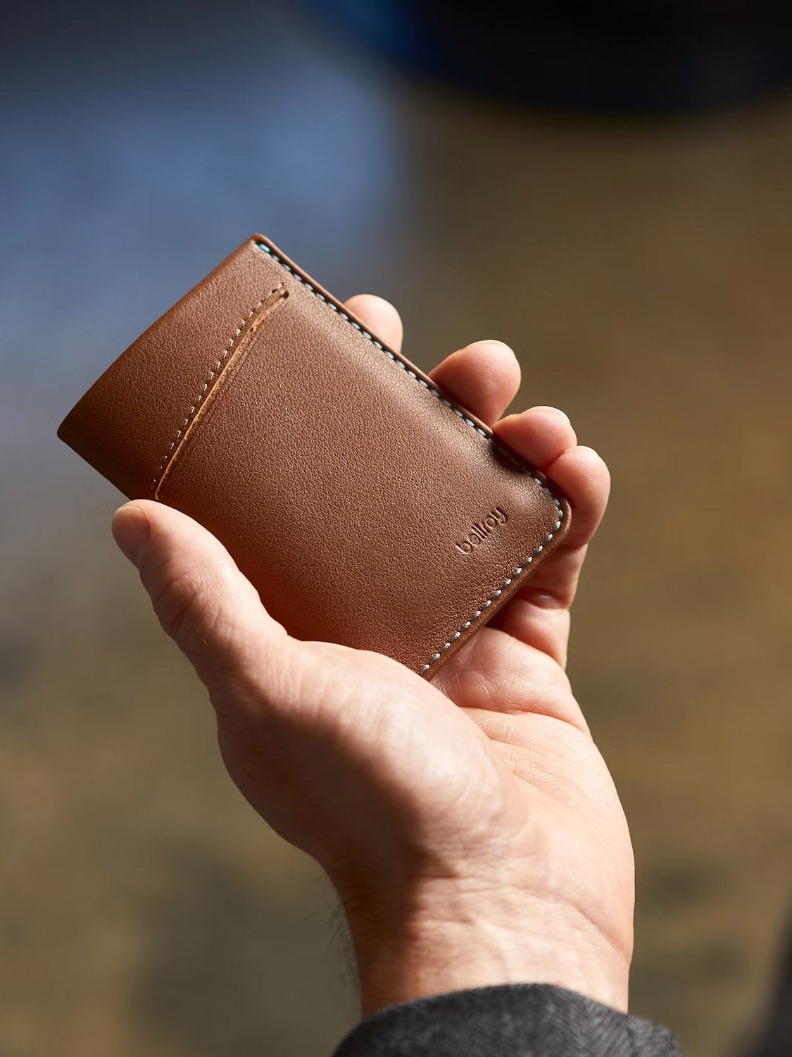 A person holds a Bellroy Card Sleeve Wallet made of brown leather with visible stitching and a small front pocket. "Bellroy" is embossed on the lower right corner, and the softly blurred background highlights its minimalist design.