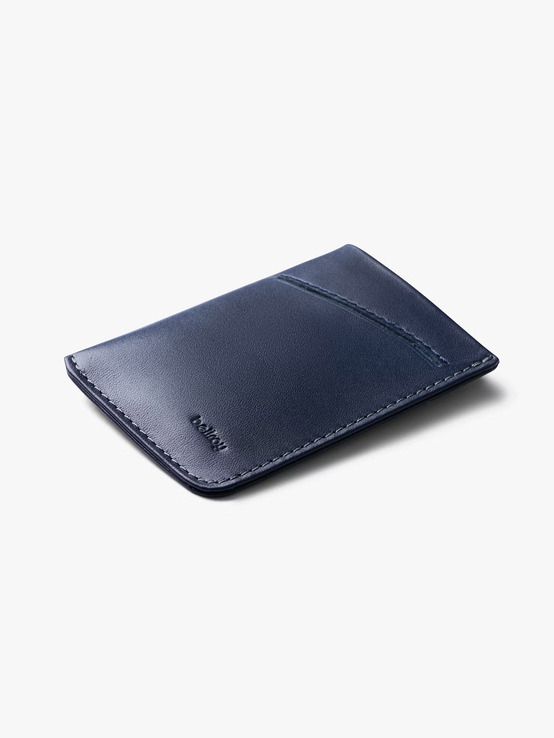 The Bellroy Card Sleeve Wallet is a slim, navy blue minimalist leather wallet with a single angled card slot, visible edge stitching, and an embossed logo on the bottom left. Shown lying flat on a light gray surface.