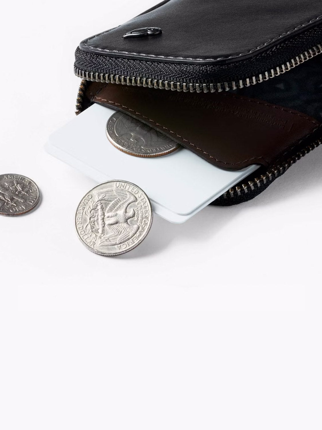 A Bellroy Card Pocket Wallet in black leather sits slightly open, showing coins and cards inside. Two quarters and a card peek out, while another quarter rests outside on a white surface.