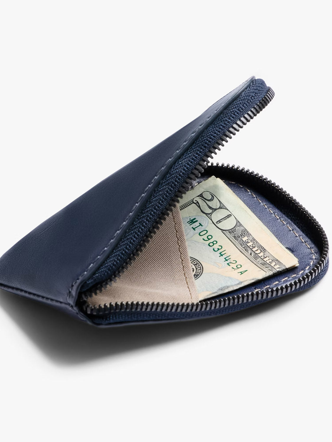 A Bellroy Card Pocket Wallet in navy leather with a zippered closure sits partially open, revealing a folded twenty-dollar bill, coin pouch, and beige contents inside, all set against a white background.