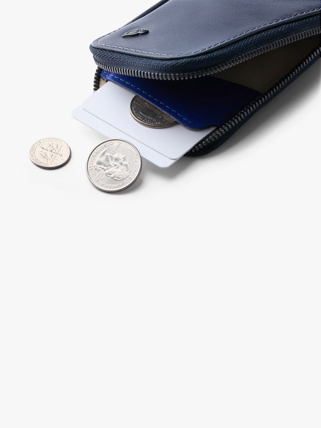 The Bellroy Card Pocket Wallet in navy blue leather is partially open, showing cards and coins inside. Three silver coins are on a white surface in front of this versatile wallet with a zippered coin pouch.