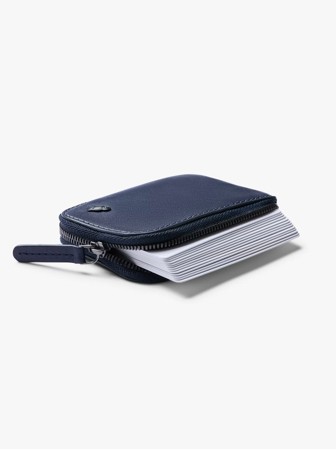 The Bellroy Card Pocket Wallet, a compact navy blue leather accessory with a zippered closure, is slightly open to reveal white cards inside. It is showcased against a plain white background.