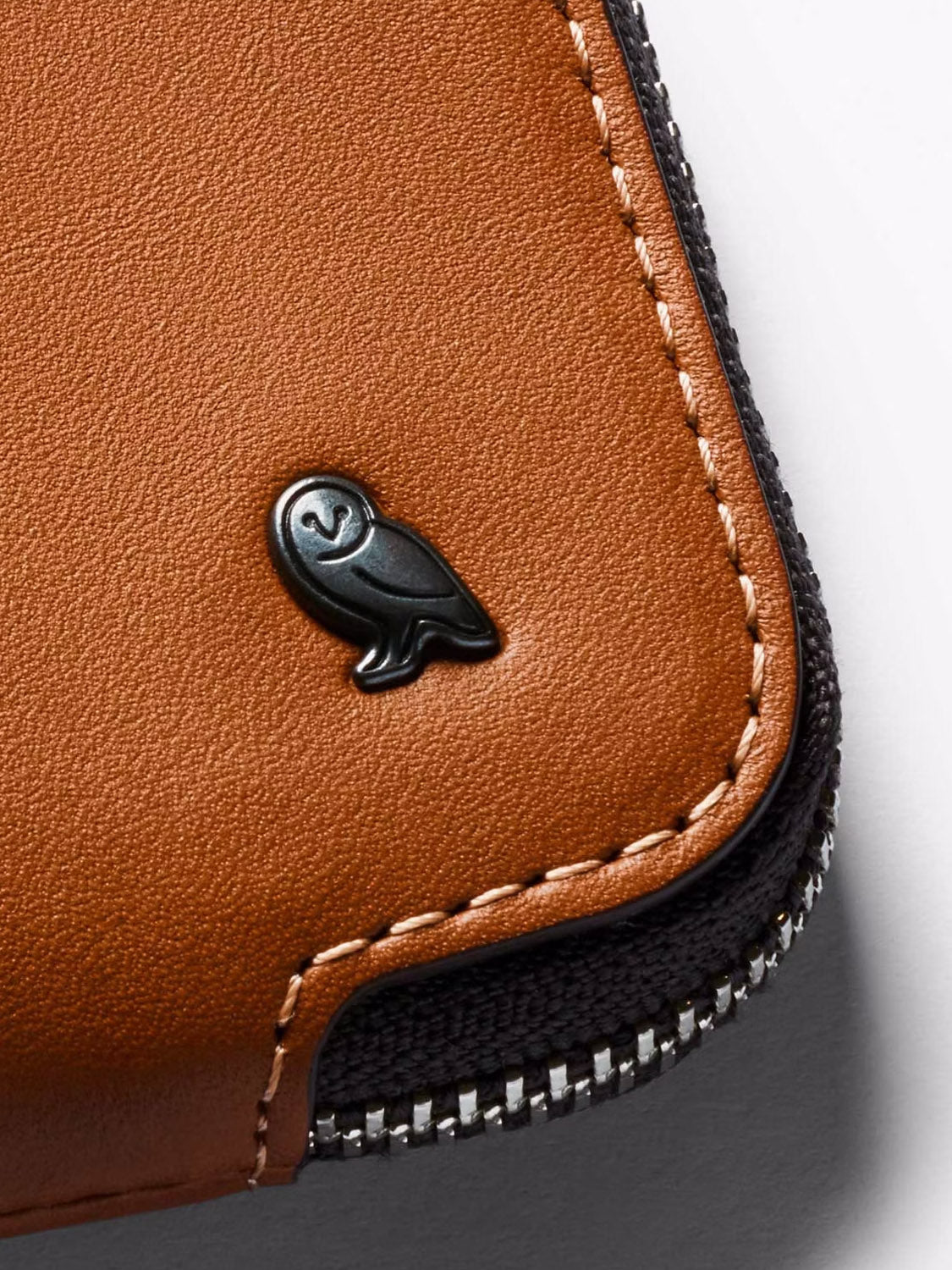 Close-up of the Bellroy Card Pocket Wallet in brown leather, featuring white stitching, a small black owl logo, black fabric trim on the edge, and a visible metal zipper.
