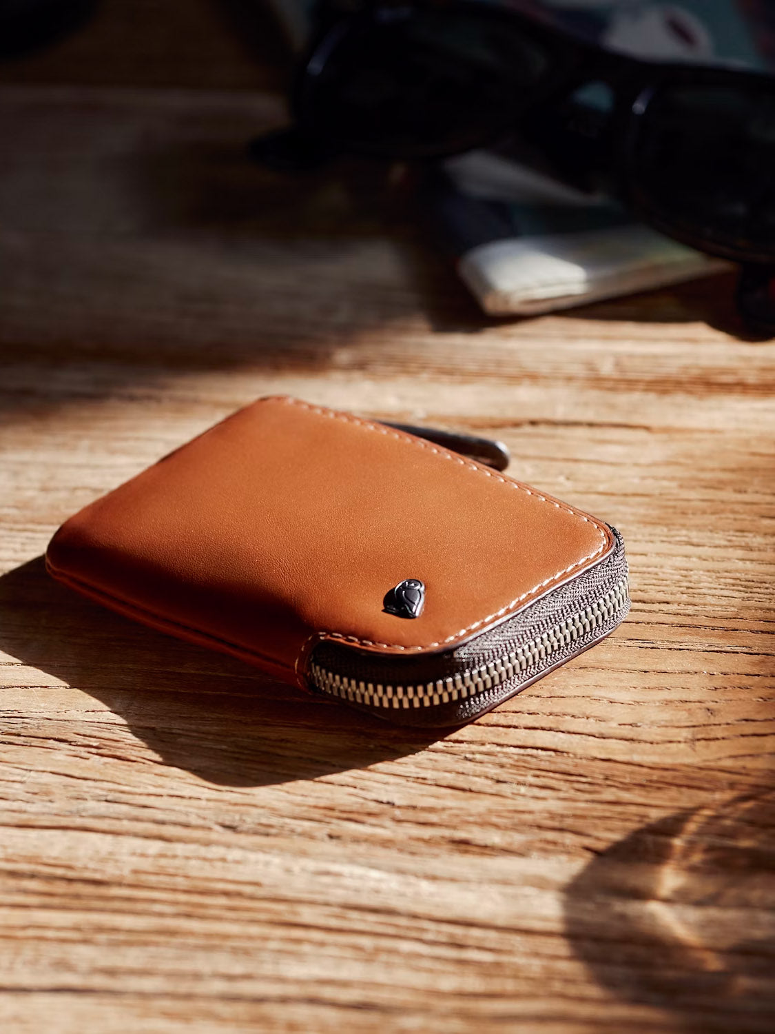 The Bellroy Card Pocket Wallet, a brown leather accessory with a zipper and card holder compartment, rests on a wooden surface. Sunglasses and other blurred items can be seen in the background.