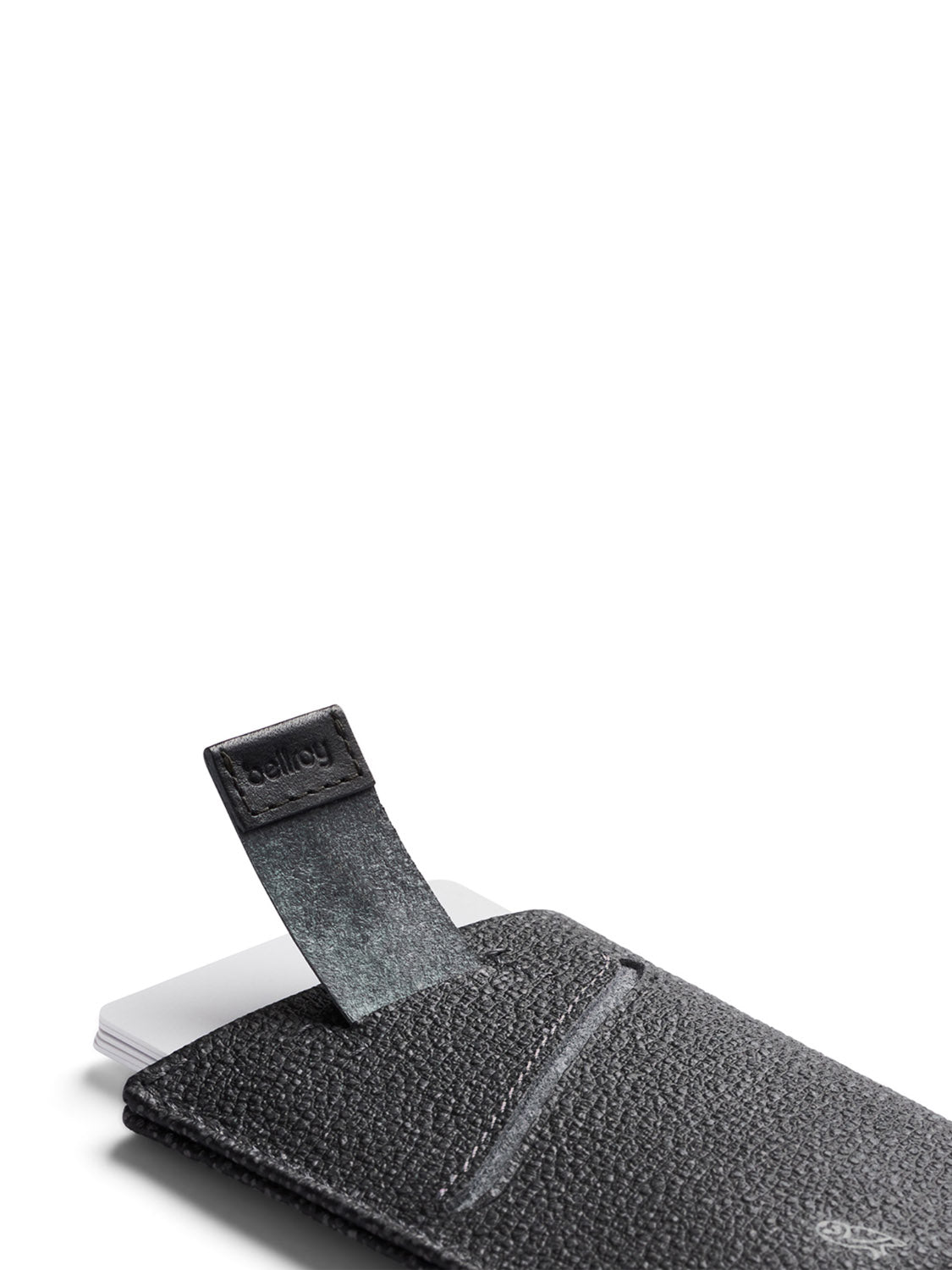 A close-up of the Bellroy Card Sleeve Wallet in black textured leather shows a small tab with "bellroy" embossed, partially pulled out to reveal the edge of a white card inside against a plain white background.