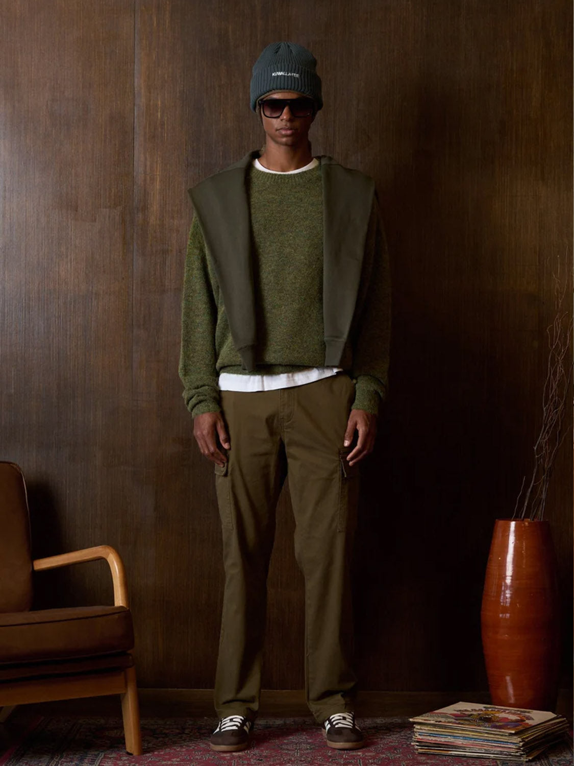 A person stands indoors against a wooden wall, wearing a green beanie, dark sunglasses, green sweater over a white shirt, and Kuwalla Loose Cargo Trouser. Black sneakers complete the look. A chair and stacked magazines are nearby.