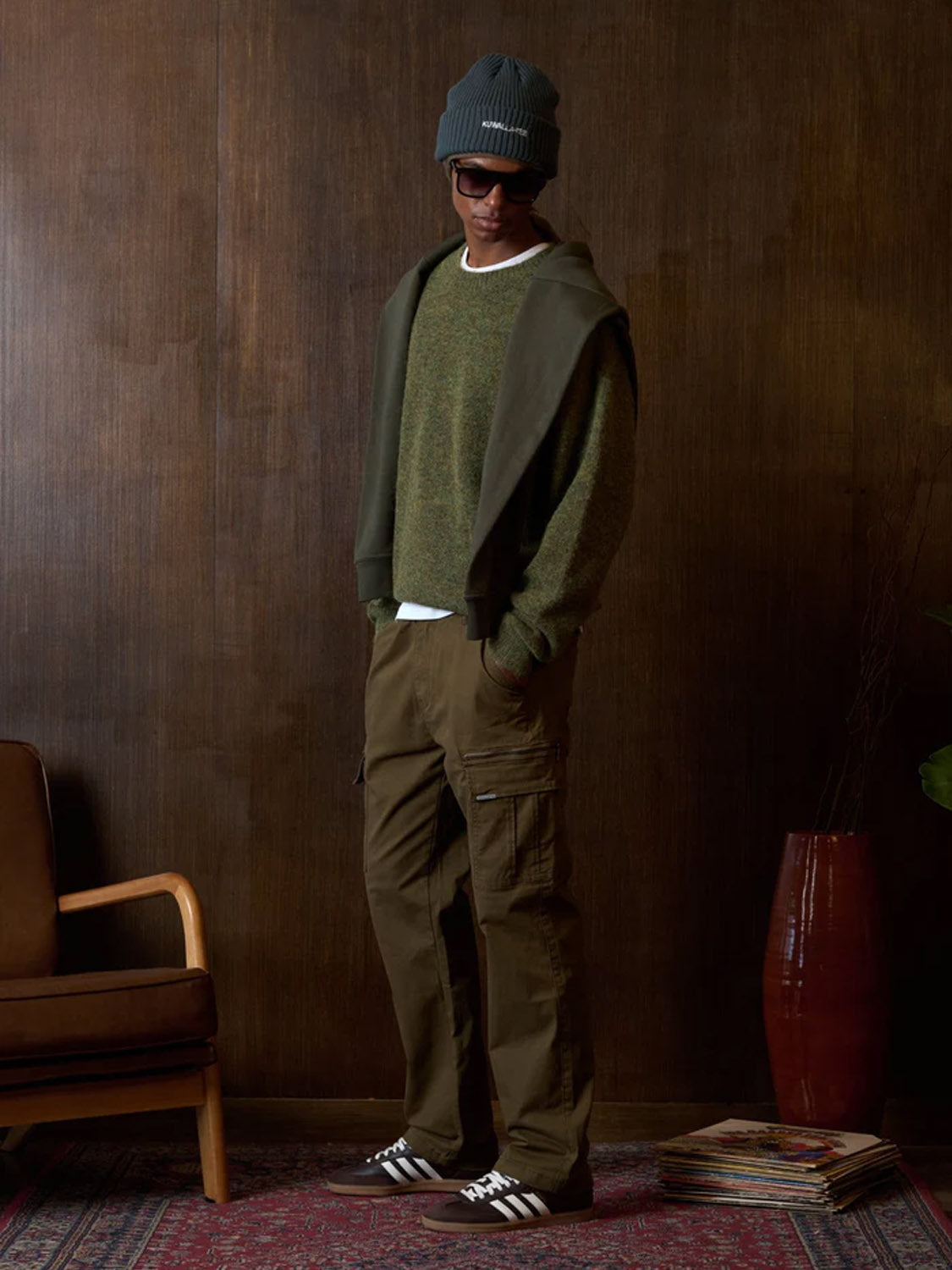 Indoors, someone wears a green beanie, dark sunglasses, an olive cotton sweater with a matching sweatshirt over the shoulders, Kuwalla Loose Cargo Trousers, brown Adidas sneakers, and stands before wooden furniture.