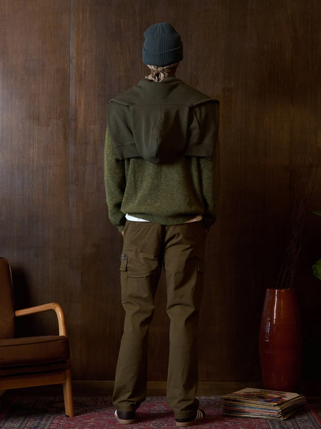 Indoors, a person faces a dark wooden wall wearing the Kuwalla Loose Cargo Trouser in brown with side pockets, a gray beanie, green sweater, jacket over shoulders, and sneakers. A chair, large red vase, and magazines are nearby.