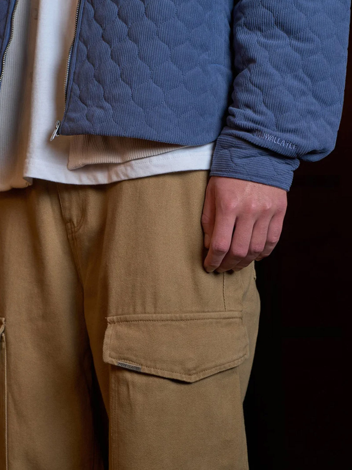 A person wears Kuwalla Cargo Trouser in a relaxed fit with a white shirt and light blue quilted jacket, their right hand at their side, set against a dark background.