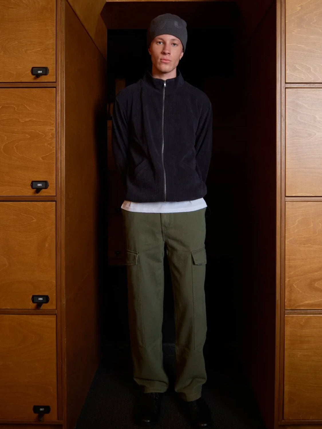 A person stands indoors between wooden cabinets, wearing a gray beanie, dark zip-up jacket, white shirt, and Kuwalla Cargo Trouser in a classic cargo style, looking at the camera with hands behind their back.