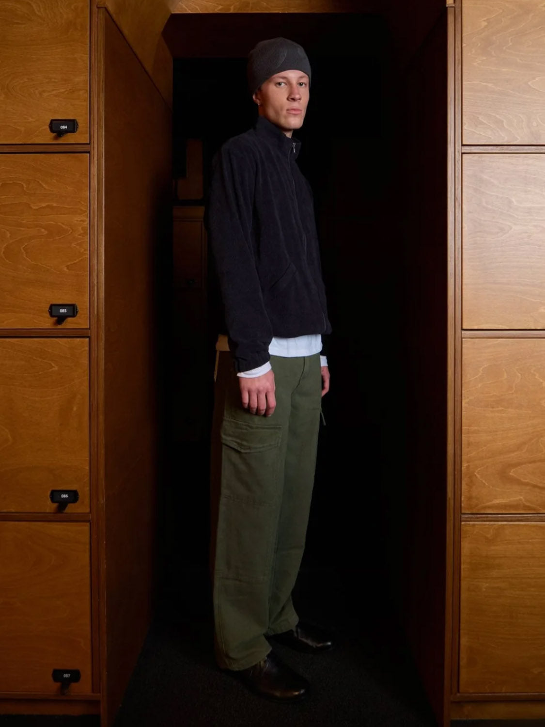 A person wears Kuwalla Cargo Trouser, a gray beanie, a dark zip-up jacket, and a white shirt while standing between wooden lockers in a dimly lit hallway, looking at the camera.